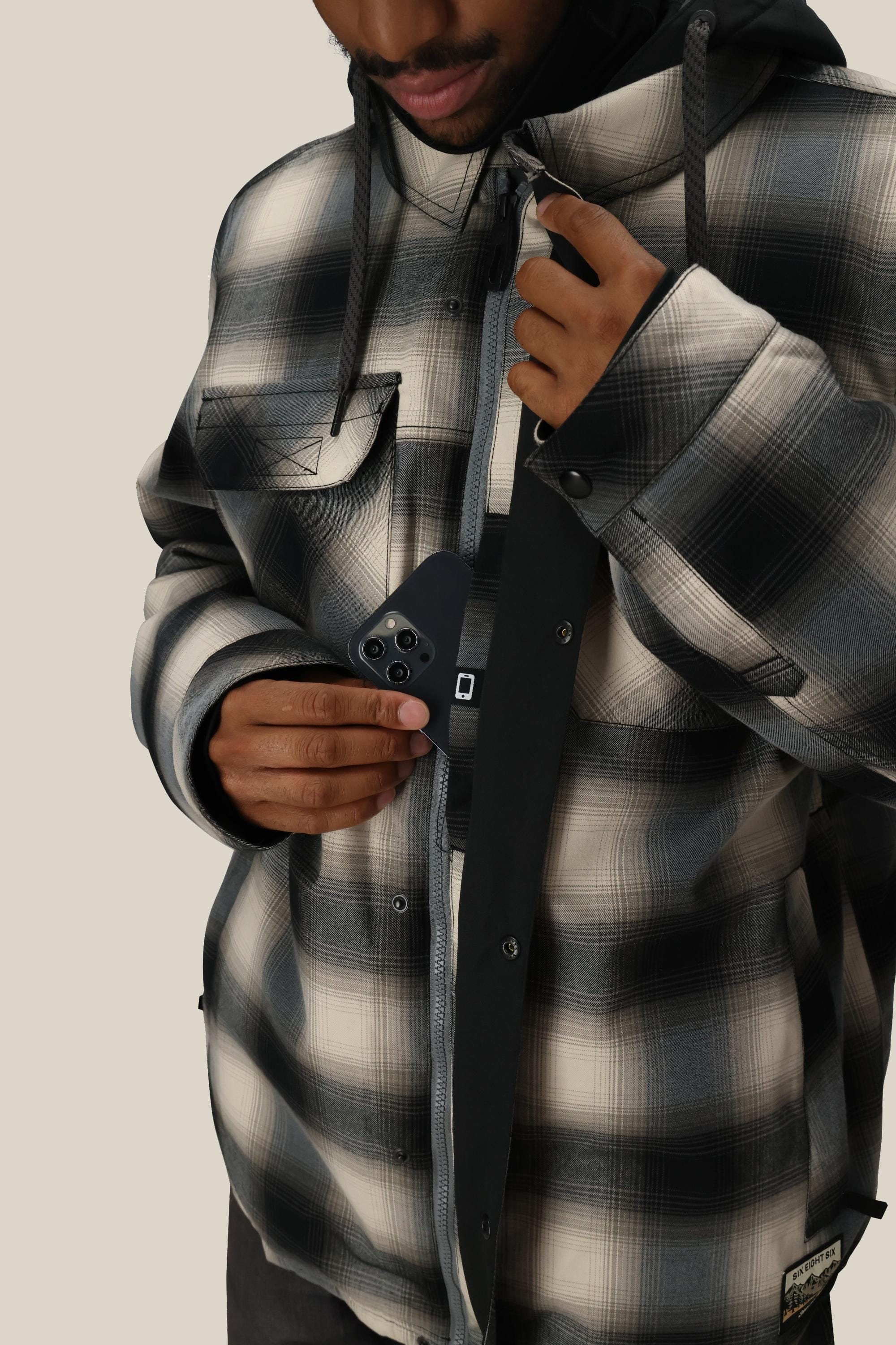 A person wearing the 686 Mens Woodland Insulated Jacket in black and white is unzipping it and placing a black smartphone into the inner chest pocket, against a plain beige background.
