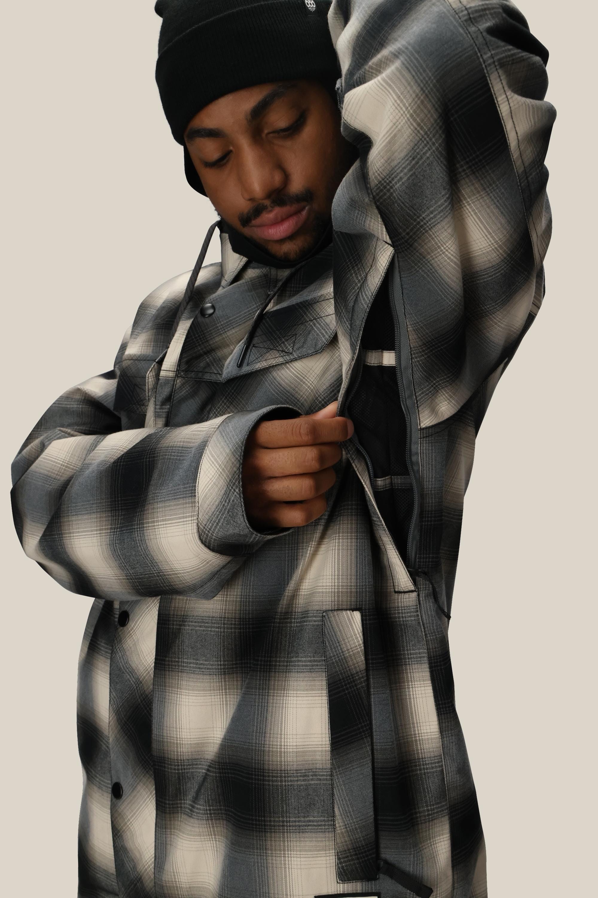 A person wearing a black beanie and the 686 Mens Woodland Insulated Jacket unzips the side ventilation, looking down with a neutral expression against a plain background.