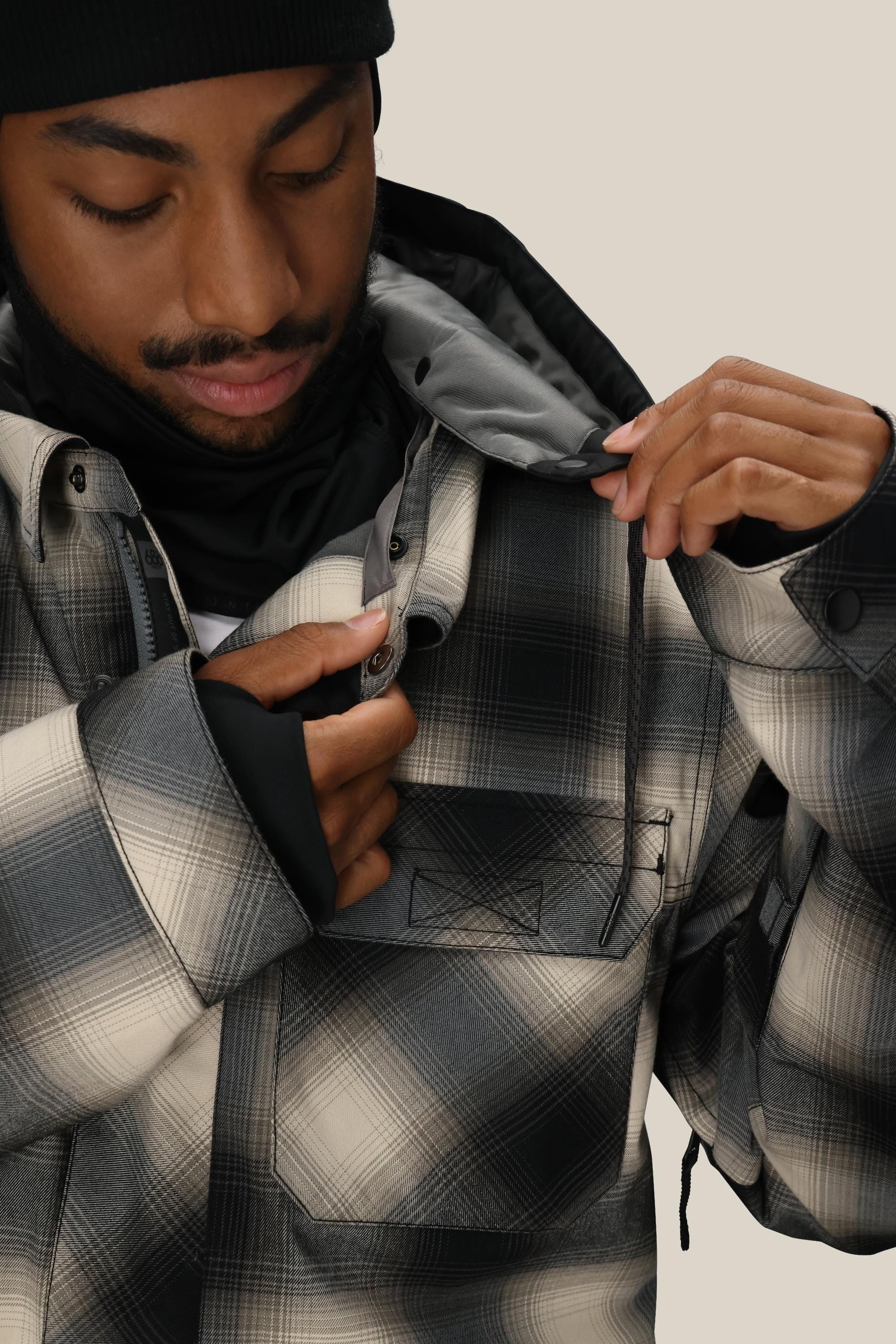 A person wearing a black beanie and the 686 Mens Woodland Insulated Jacket in black-and-grey fastens the top button while looking down with a neutral expression.
