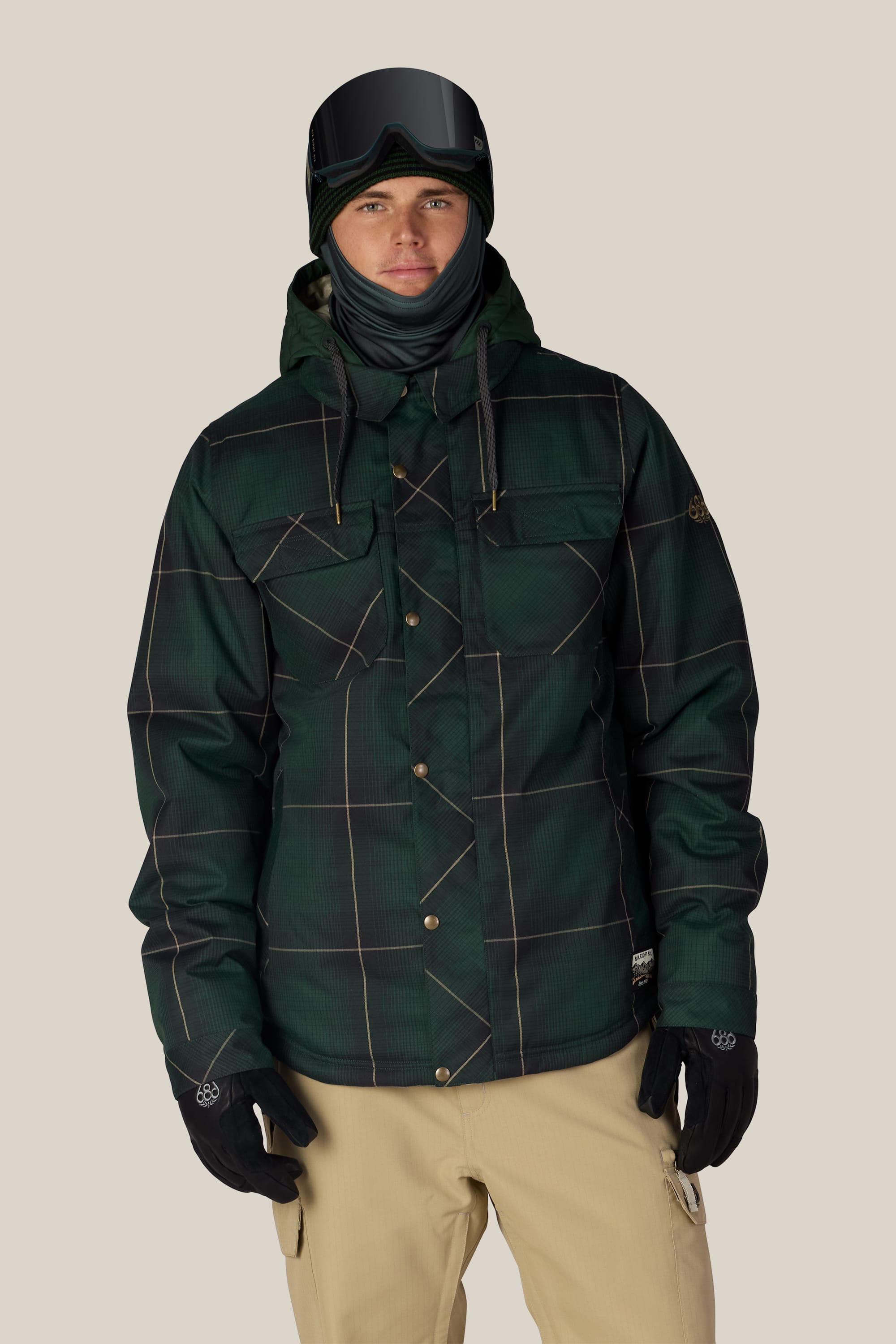 Wearing the 686 Men’s Woodland™ Insulated Jacket in dark green plaid, along with beige pants, black gloves, a striped neck gaiter, and ski goggles, a person stands against a plain light background.