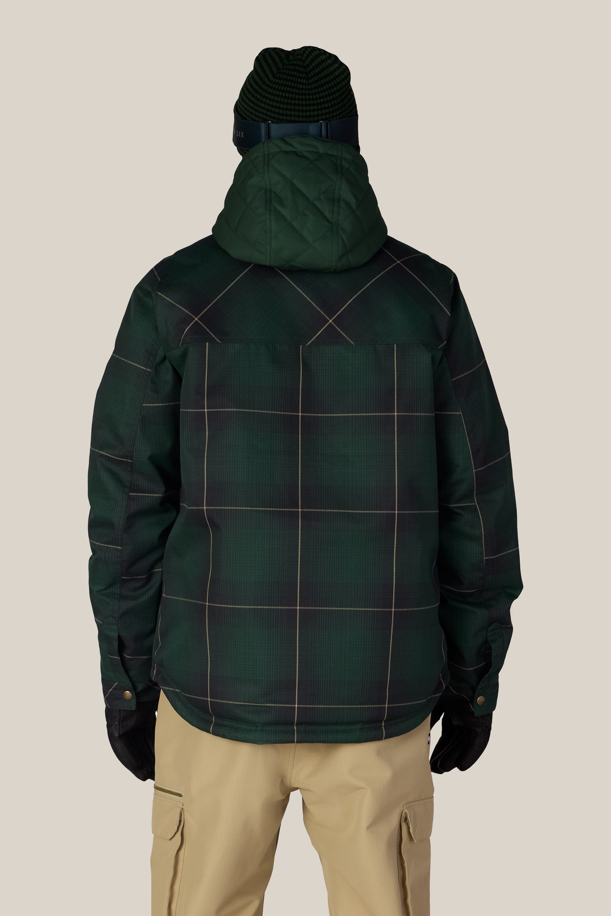A person is seen from behind against a plain light background, wearing the 686 Men’s Woodland™ Insulated Jacket in green plaid, paired with tan pants, black gloves, and a green knit beanie.