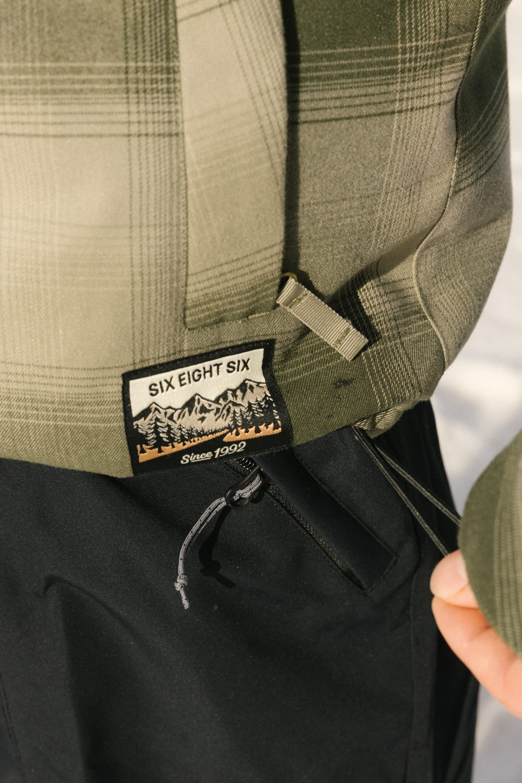 Close-up of the 686 Mens Woodland Insulated Jacket in green plaid polyester, featuring a 686 patch above a mountain scene and Since 1992. Paired with black pants; a hand is visible at the edge.