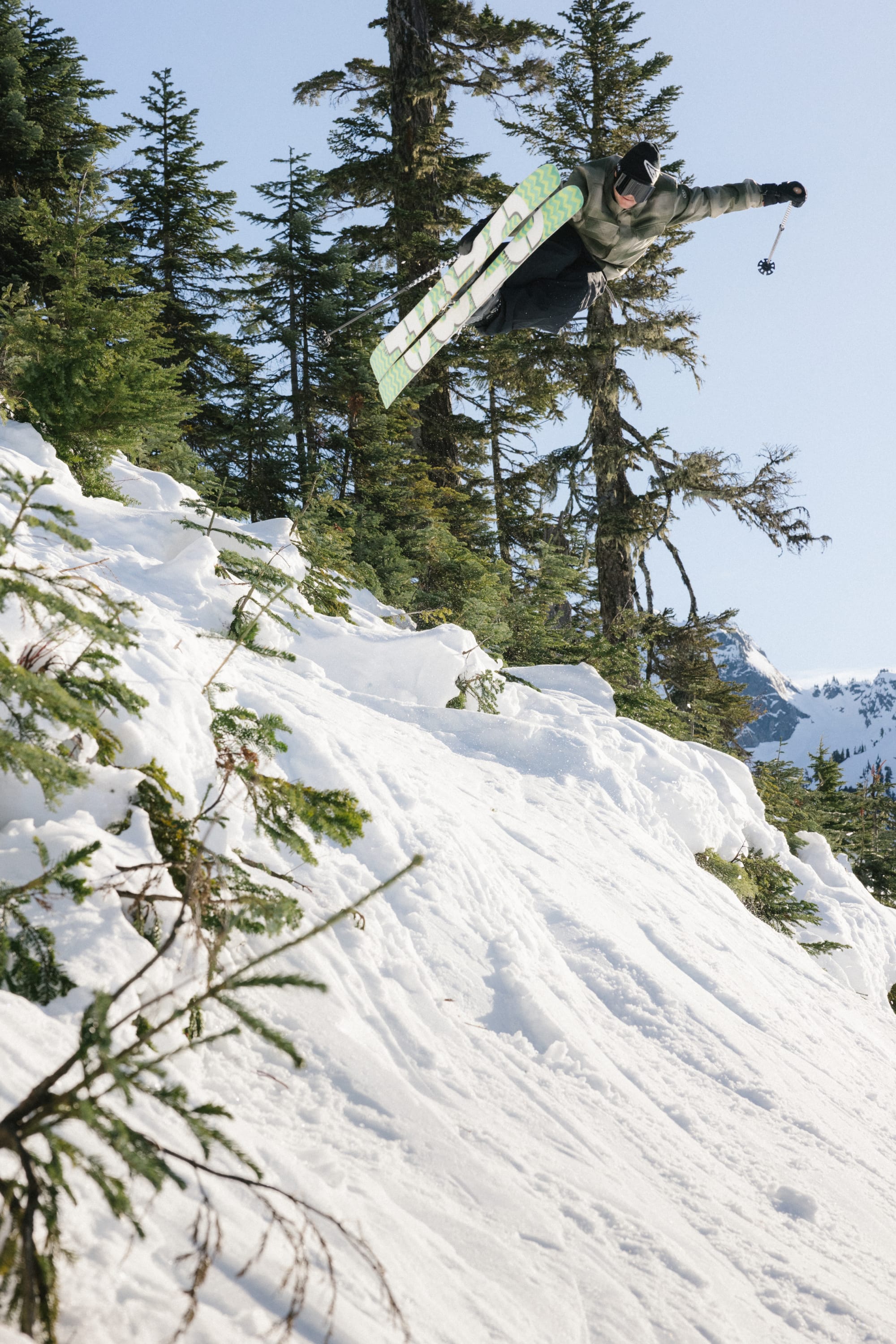 A snowboarder in the 686 Mens Woodland Insulated Jacket soars above a snowy slope, surrounded by pine trees, under a sunny blue sky.