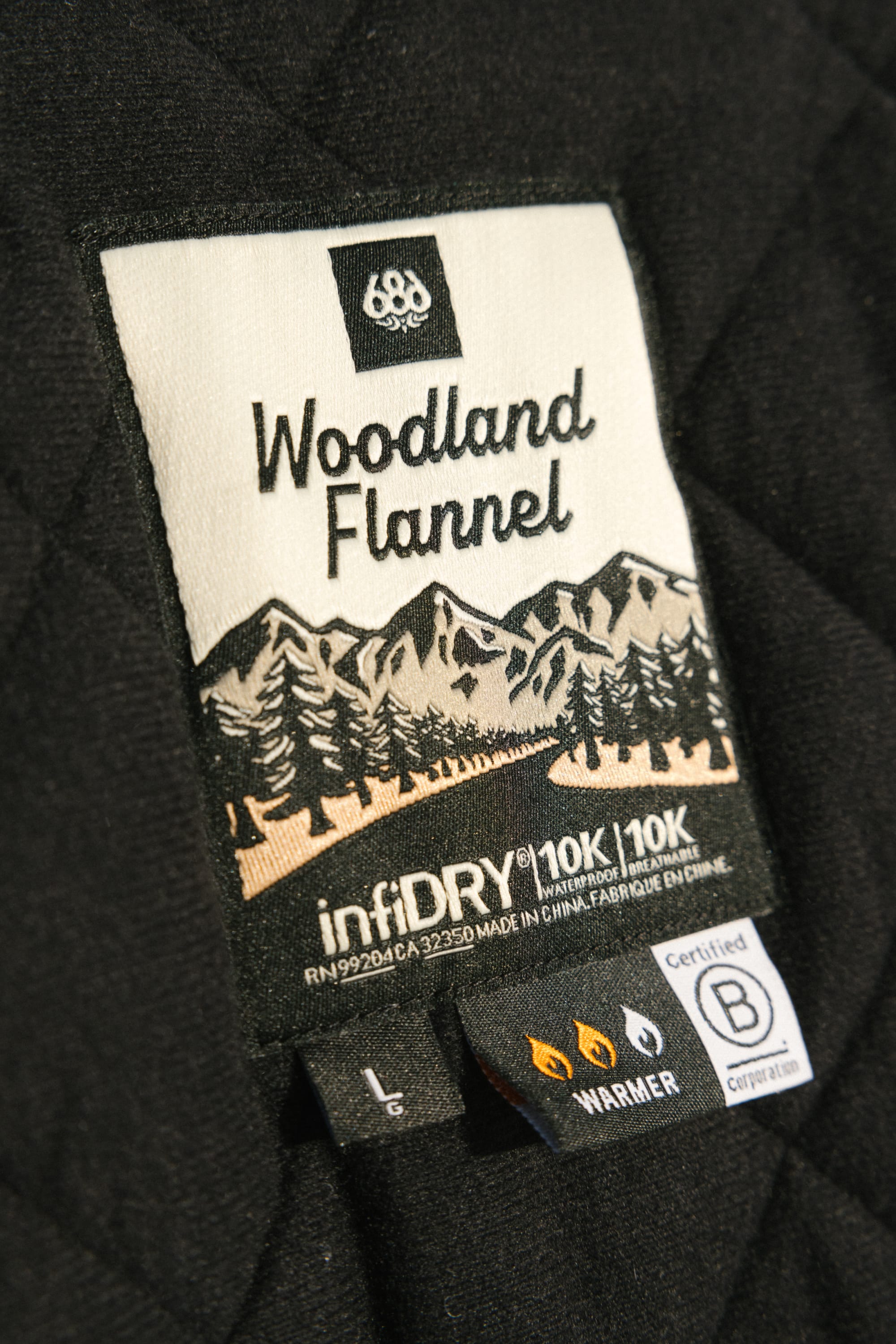 Clothing tag on a black quilted 686 Mens Woodland Insulated Jacket, featuring mountain and trees graphic, infiDRY, Warmer, certification icons, and the 686 brand.
