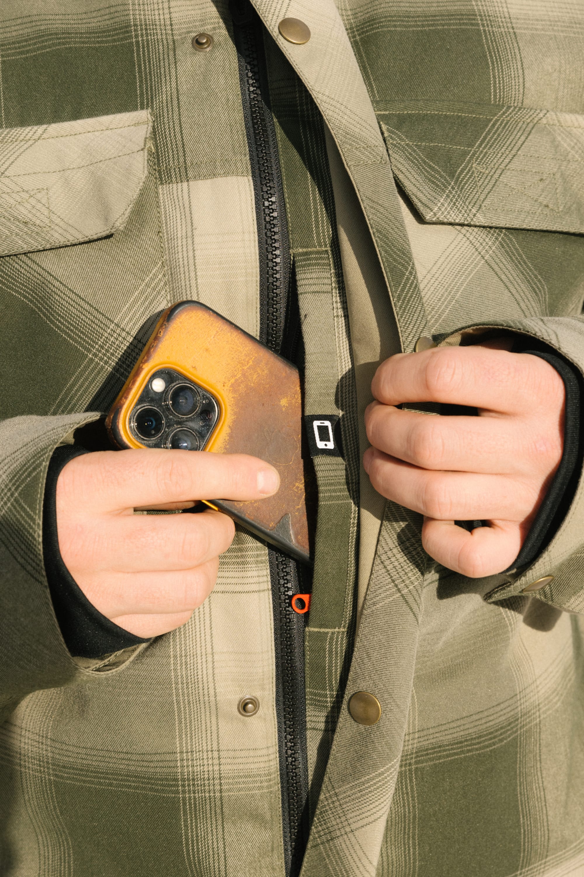 Close-up of a person in a 686 Mens Woodland Insulated Jacket, holding a phone with a brown case in their right hand and zipping up the green plaid jacket with their left.