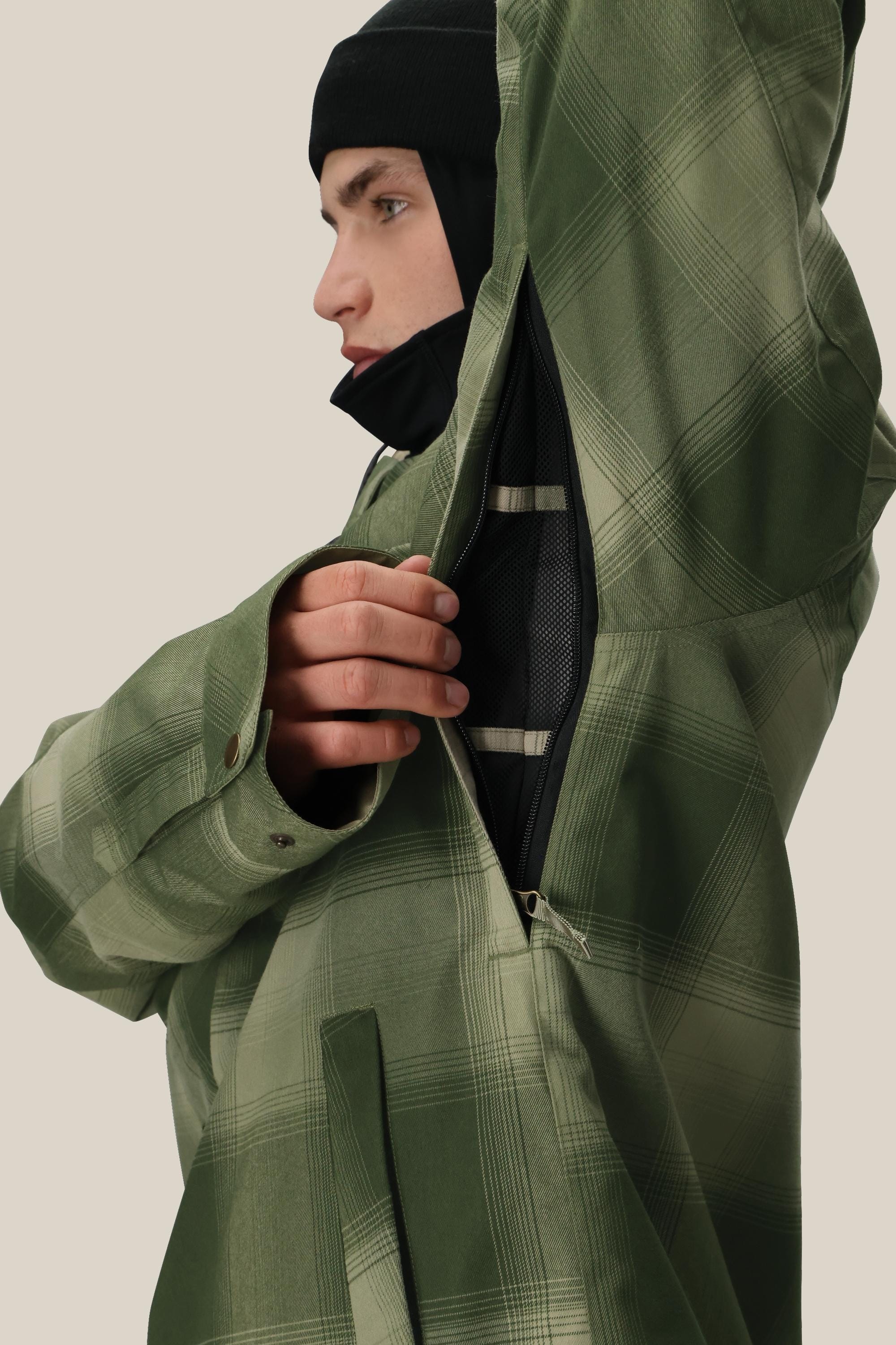 A man in a green plaid 686 Mens Woodland Insulated Jacket and black beanie opens the underarm zipper to show mesh lining for ventilation. The background is plain and light-colored.
