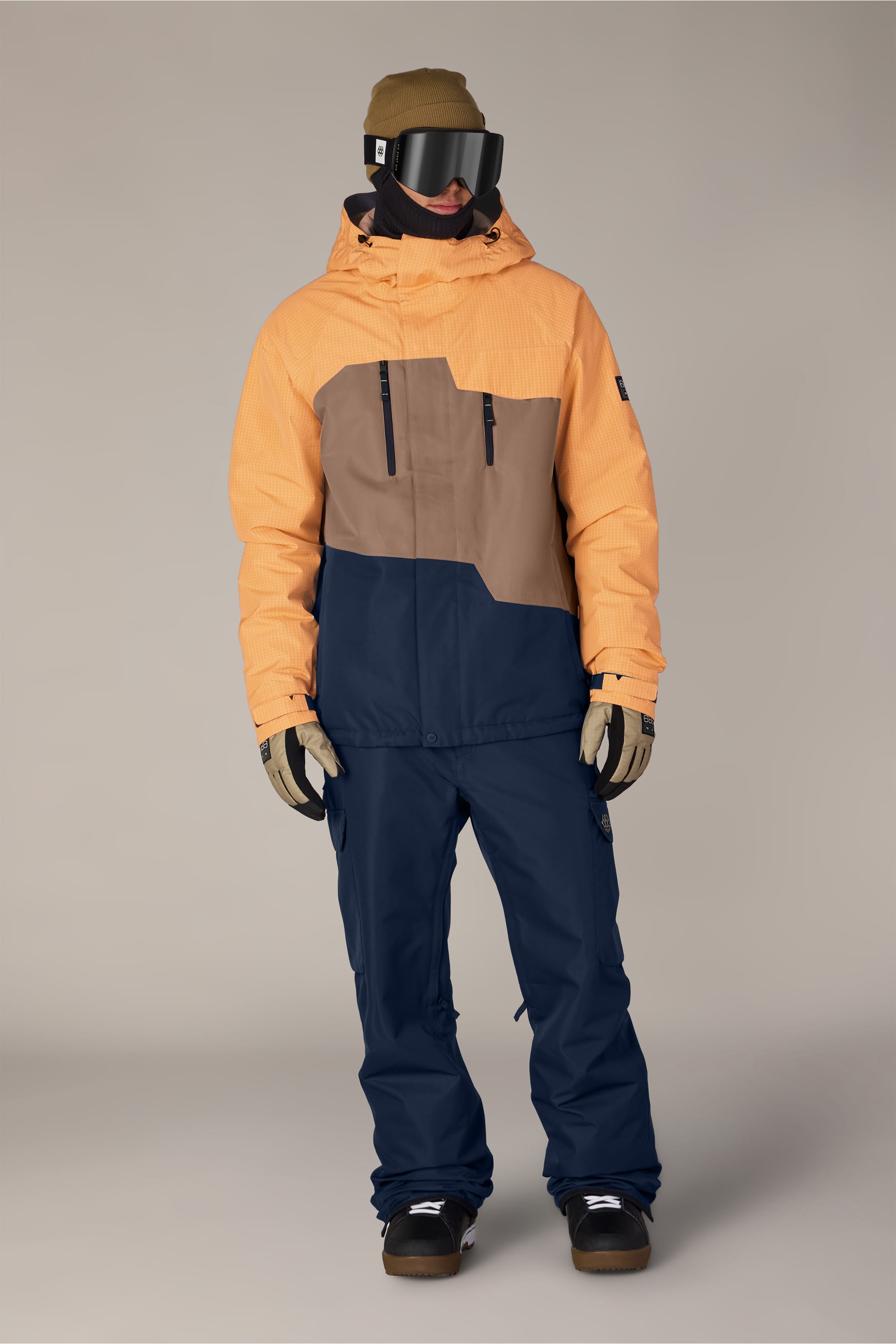 A person wearing a tan hat, black goggles, the 686 Men’s Geo™ Insulated Jacket by 686 in orange and brown, blue pants, gloves, and brown boots stands against a plain background.