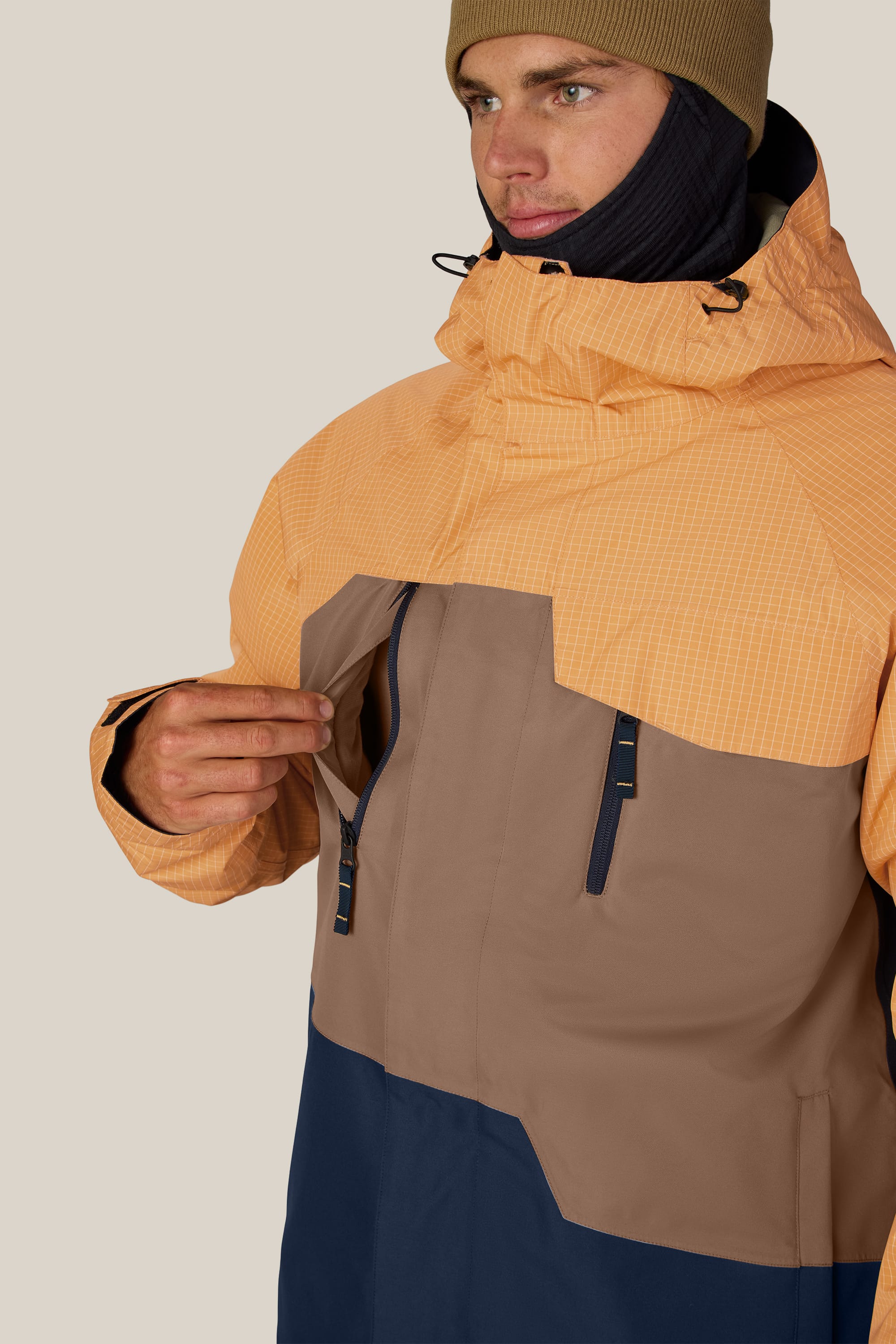 A person wearing a tan beanie and the 686 Men’s Geo™ Insulated Jacket by 686—with orange, brown, and navy color-blocked panels—demonstrates pulling the zipper on the chest pocket. The background is plain and light-colored.