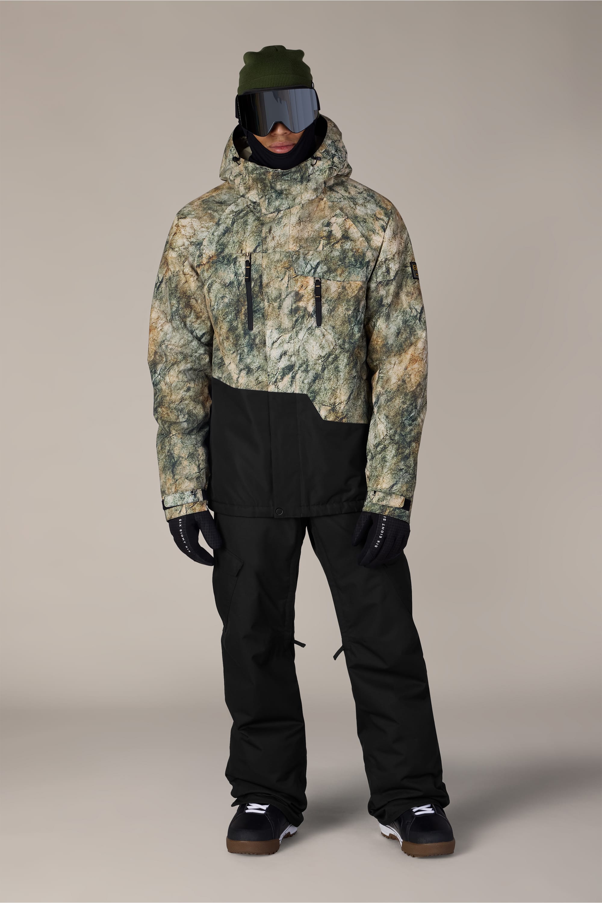 A person wearing the 686 Men’s Geo™ Insulated Jacket by 686, styled in a green and brown camo pattern, paired with black pants, black gloves, dark goggles, a green beanie, and black shoes, standing against a neutral background.