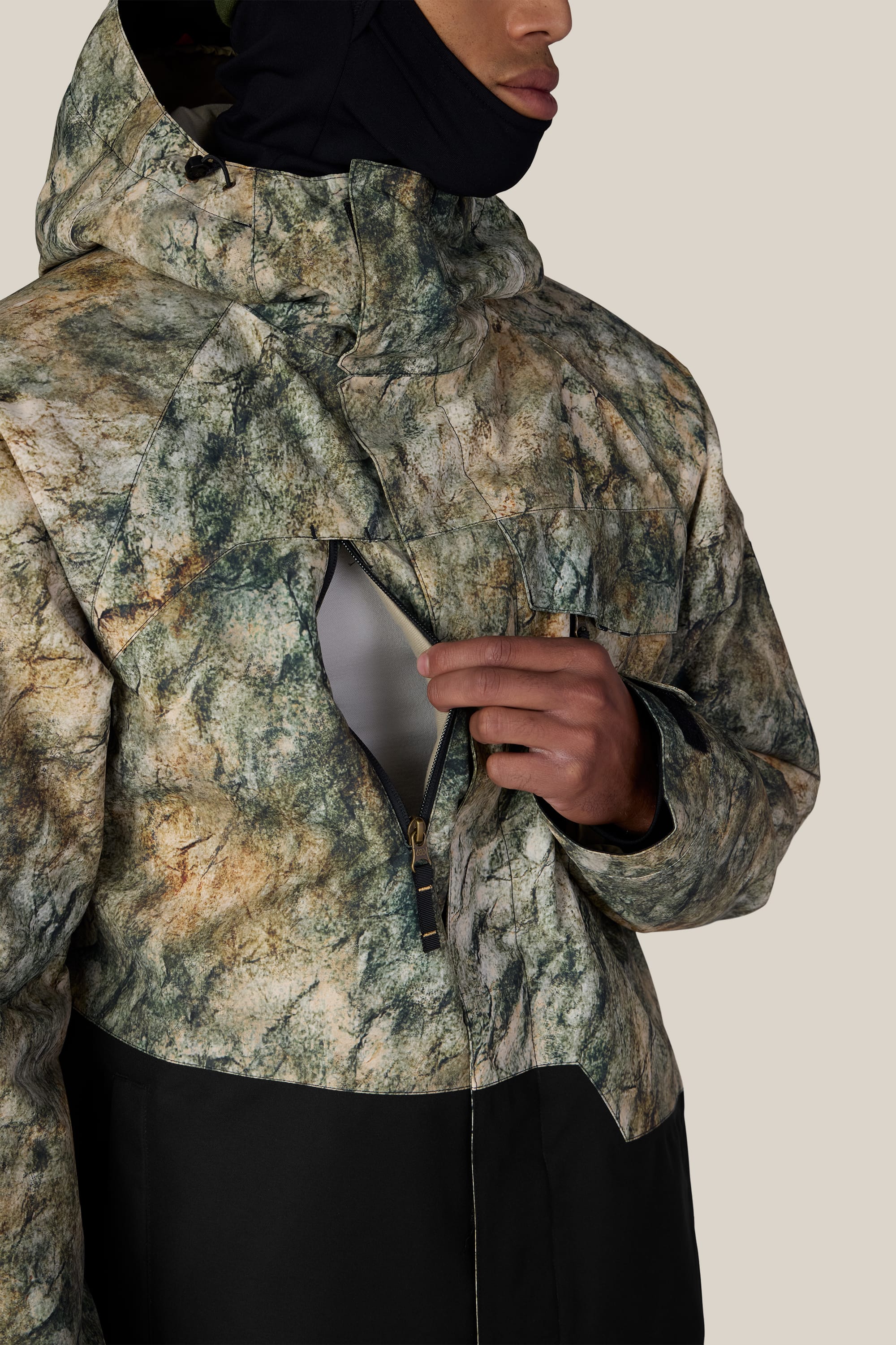 A person wearing the 686 Men’s Geo™ Insulated Jacket—a green and brown camouflage woven jacket with a black hood—un-zips the chest pocket, revealing a glimpse of the light gray inner lining.