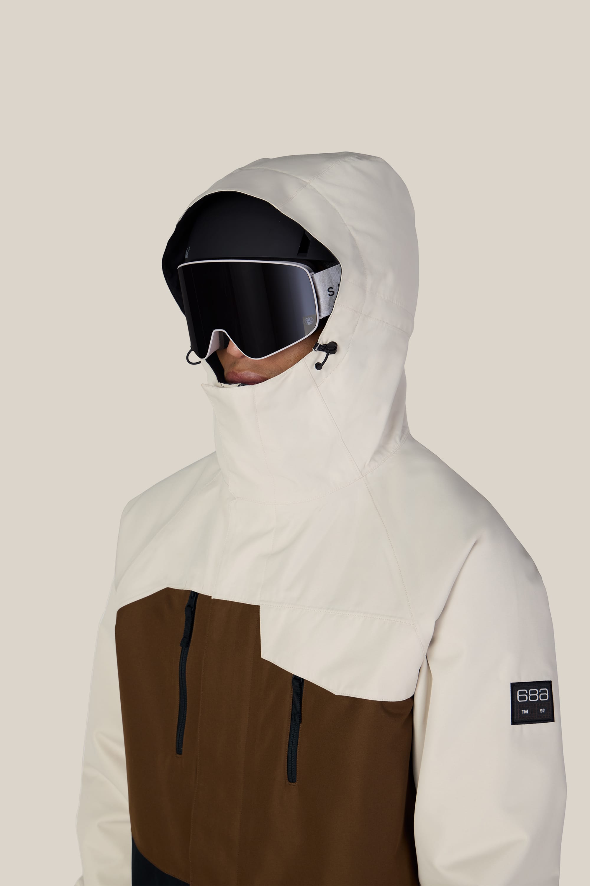 A person wearing the 686 Men’s Geo™ Insulated Jacket by 686 in a brown and cream colorway, with the hood up and large black ski goggles, faces slightly to the right against a plain light background.
