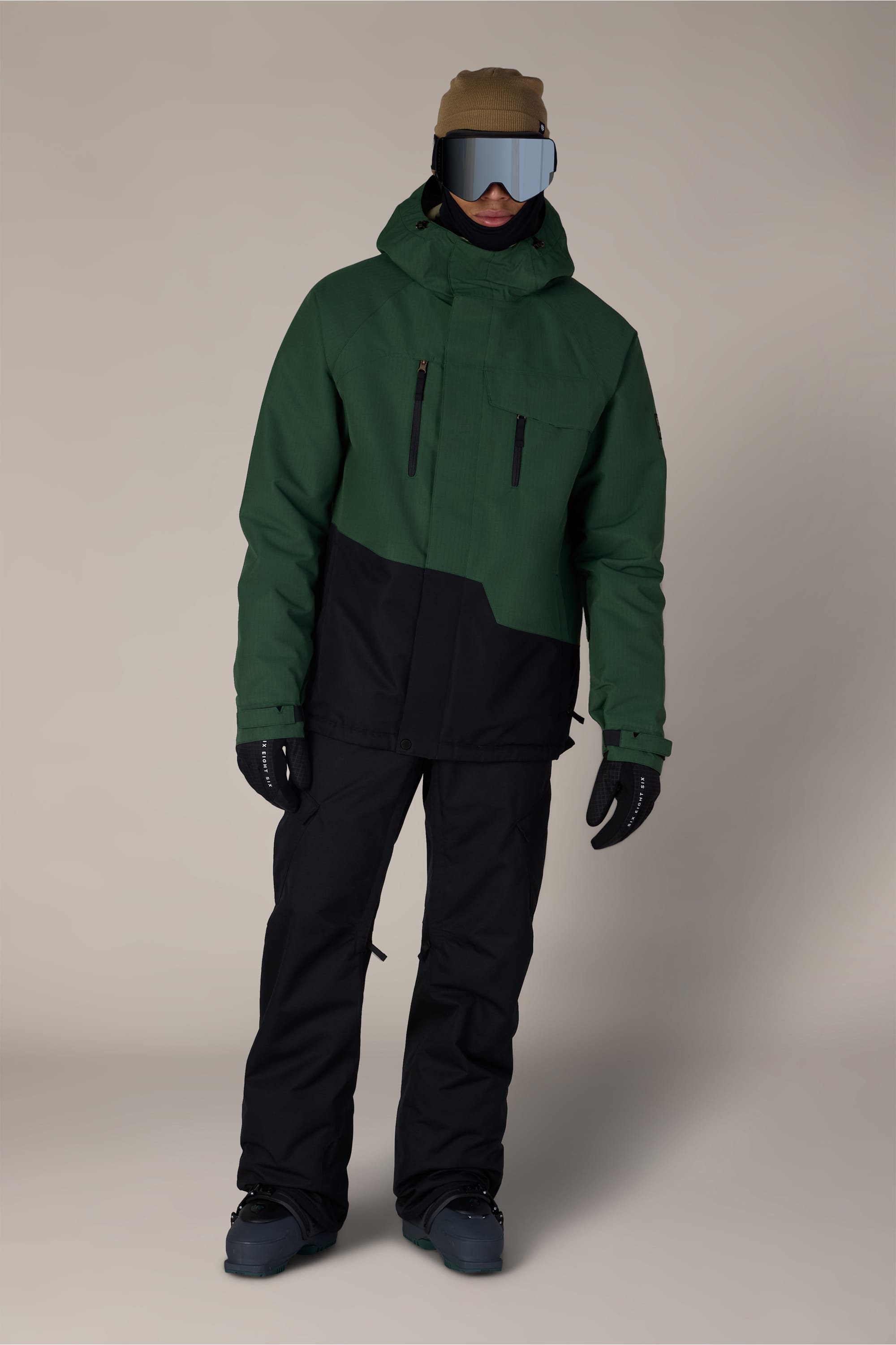 Person wearing a green and black 686 Men’s Geo™ Insulated Jacket, black snow pants, boots, gloves, a tan beanie, and mirrored ski goggles, standing against a plain light background.