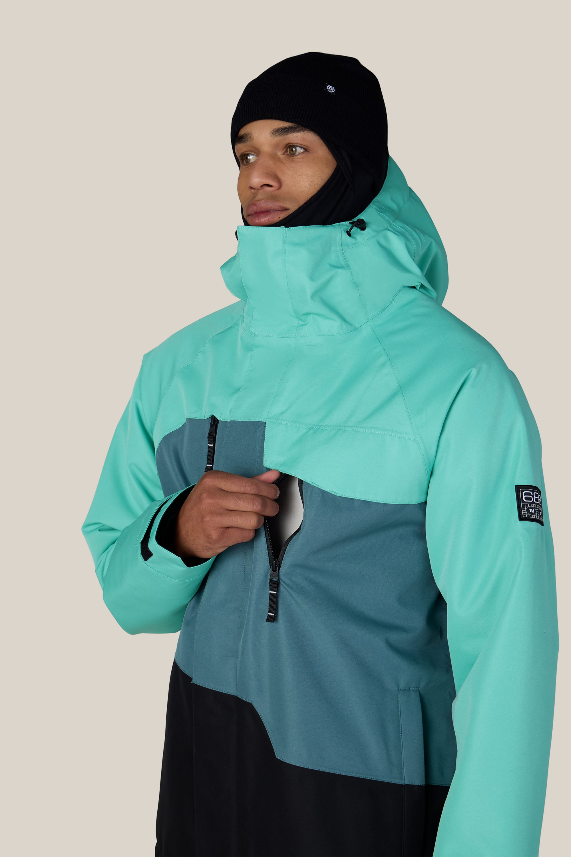 A person wearing the 686 Men’s Geo™ Insulated Jacket in teal and dark green, along with a black beanie, zips up the front while facing slightly to the right against a plain light background.