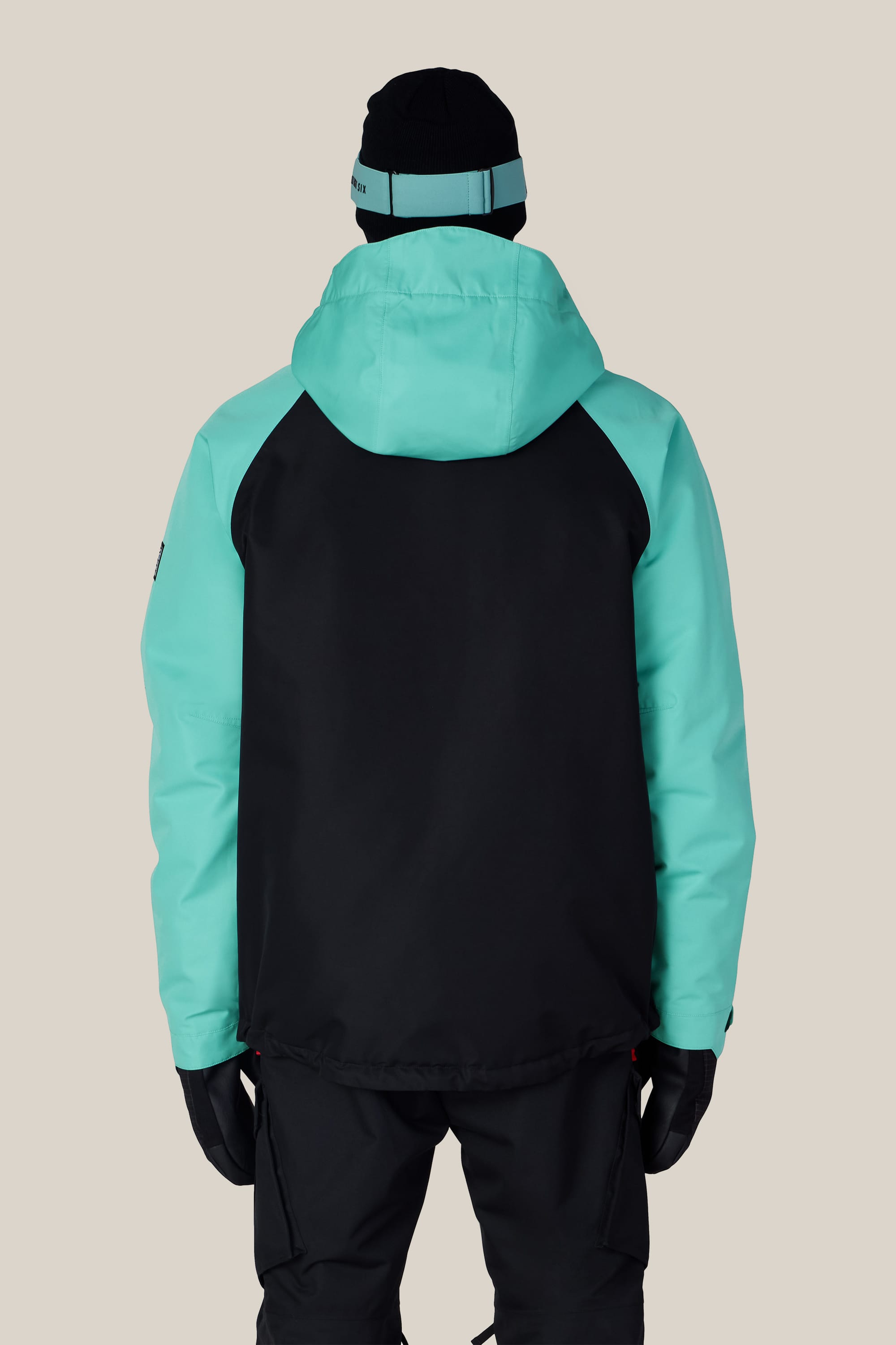 A person stands facing away against a plain beige background, wearing the 686 Men’s Geo™ Insulated Jacket in black and teal, paired with black pants, black gloves, and a black beanie featuring a teal stripe.