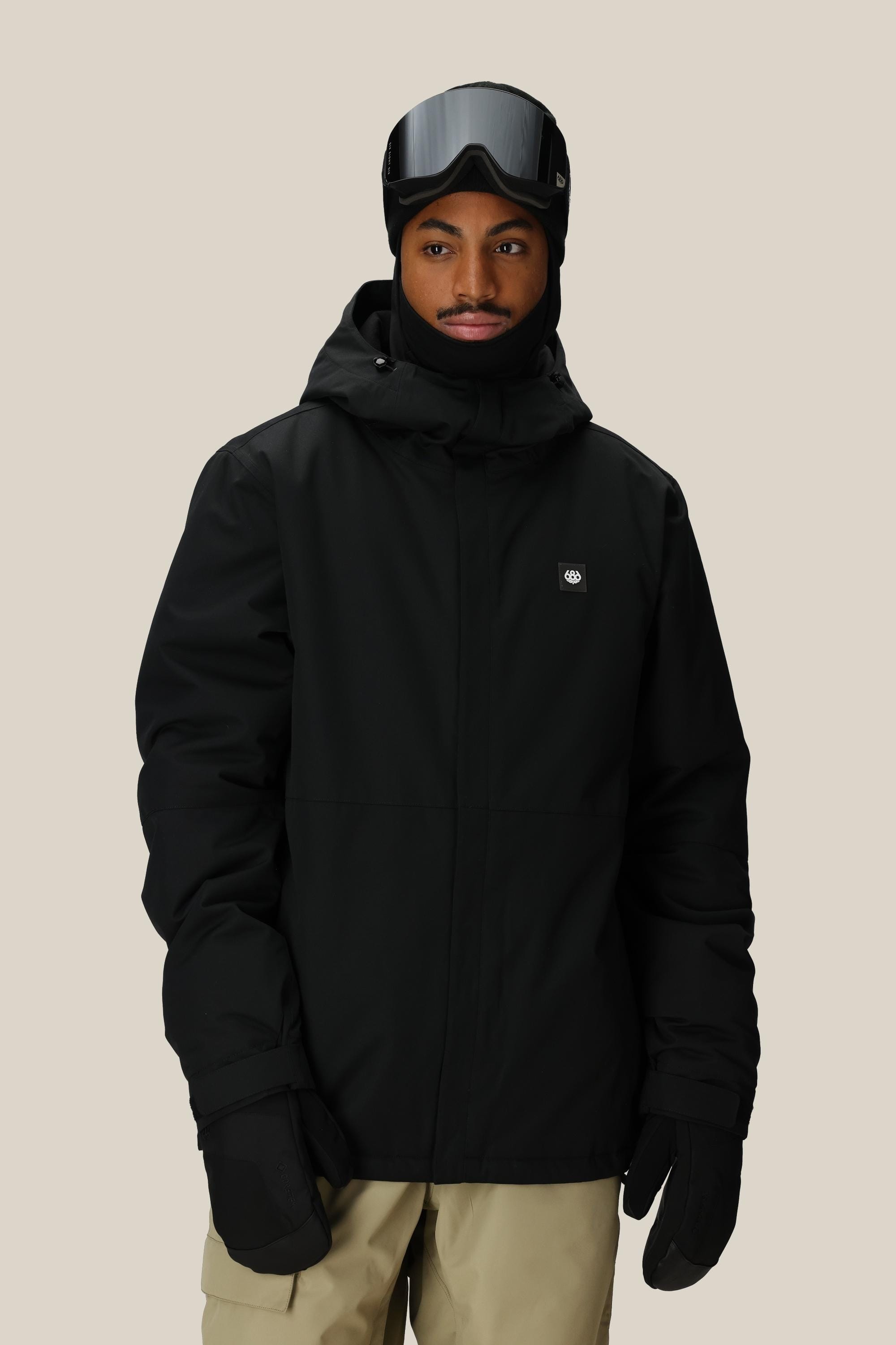 A person wearing the 686 Mens Foundation Insulated Jacket, black gloves, a black hood, ski goggles, and beige pants stands against a plain light background.