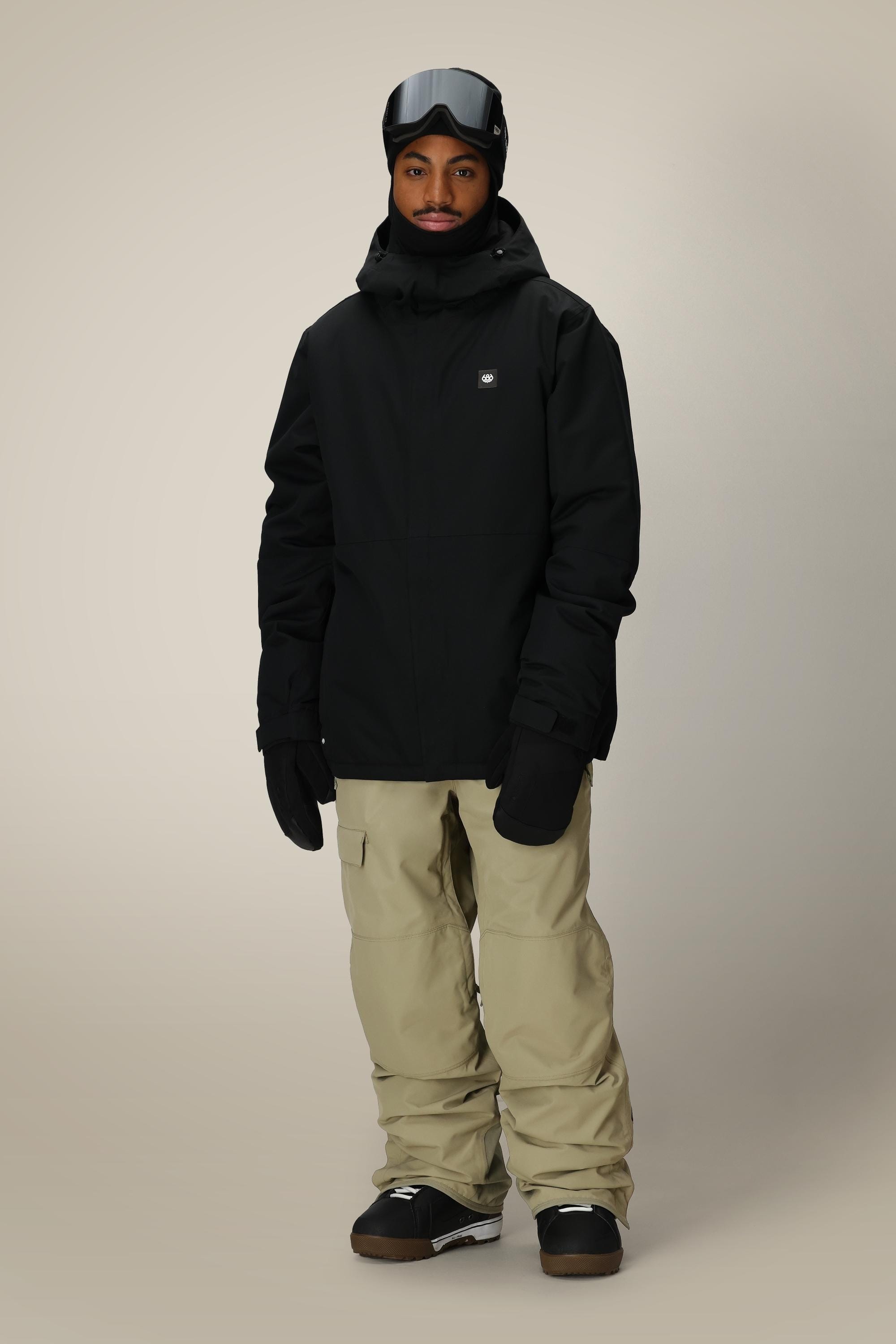 A person wears the 686 Mens Foundation Insulated Jacket with a hood, along with black gloves, black goggles on their forehead, beige snow pants, and black boots, standing against a plain light background.