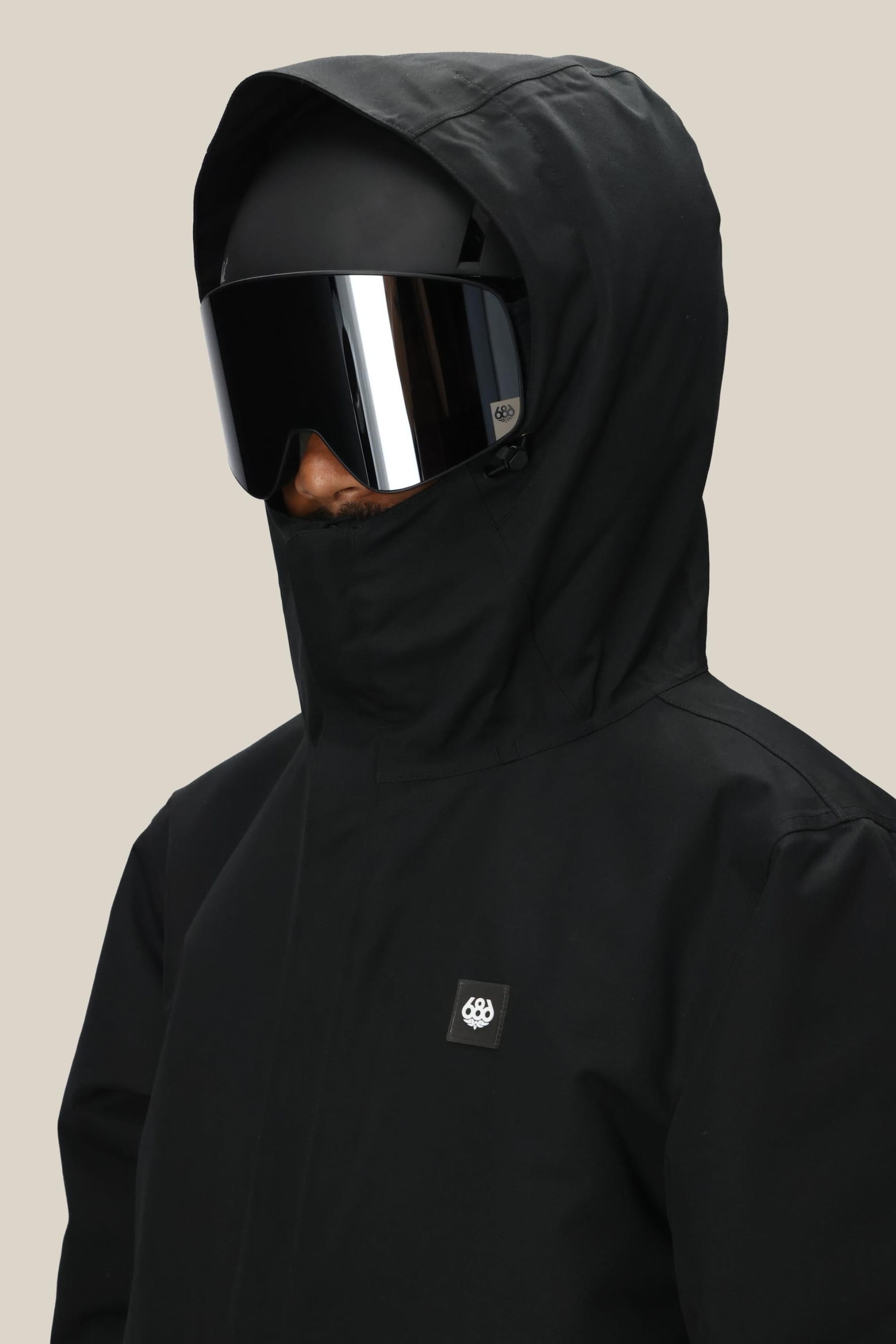 A person in a 686 Mens Foundation Insulated Jacket, black with a hood, and large reflective ski goggles faces slightly left, showing only their lower face against a plain, light background.