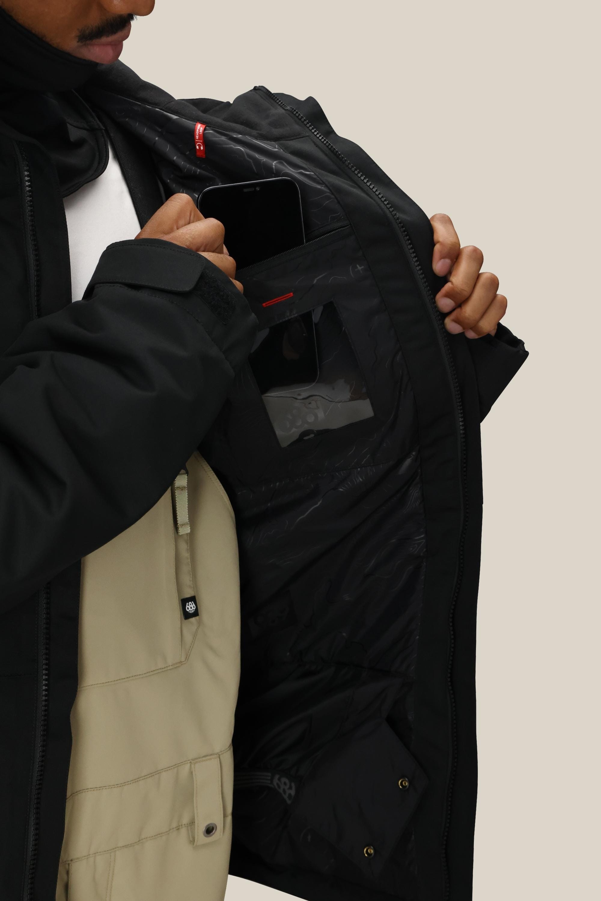 A man opens a 686 Mens Foundation Insulated Jacket, revealing inner pockets—one holding a smartphone, another with a transparent window—over a beige shirt.
