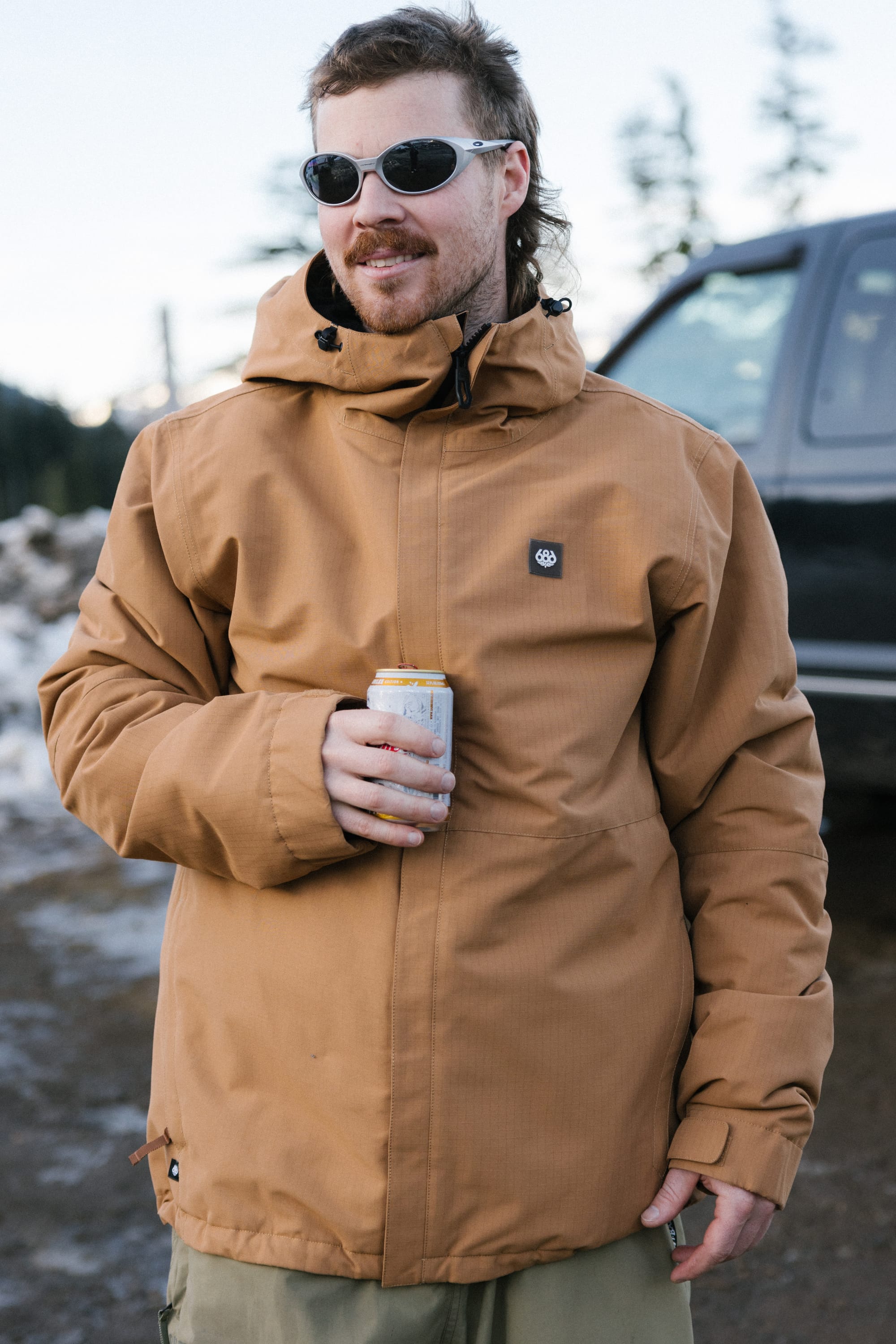 A man in the 686 Mens Foundation Insulated Jacket and sunglasses holds a beverage can outdoors beside a dark vehicle, with snow and trees in the background.