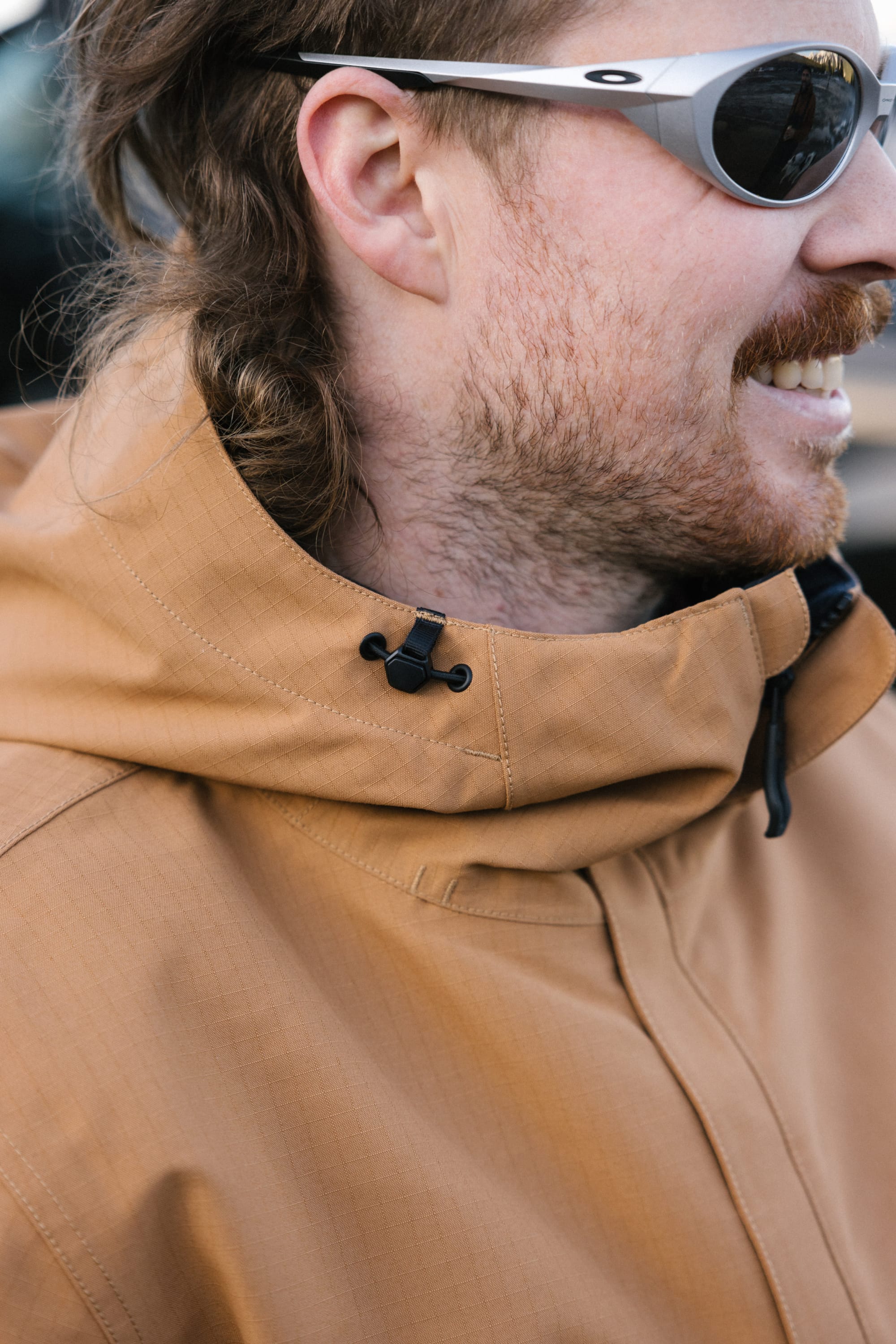 A man with light facial hair, smiling and looking to the right, wears gray sunglasses and a tan 686 Mens Foundation Insulated Jacket with a high collar.