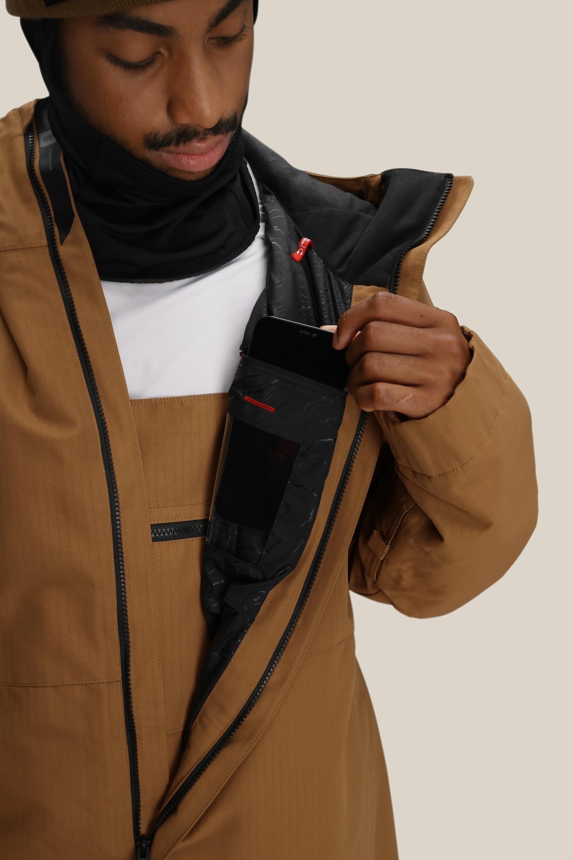 Wearing a 686 Mens Foundation Insulated Jacket in brown, a person in a black balaclava slips a smartphone into the inner zippered chest pocket, visible through the partially unzipped waterproof coat with its black lining.