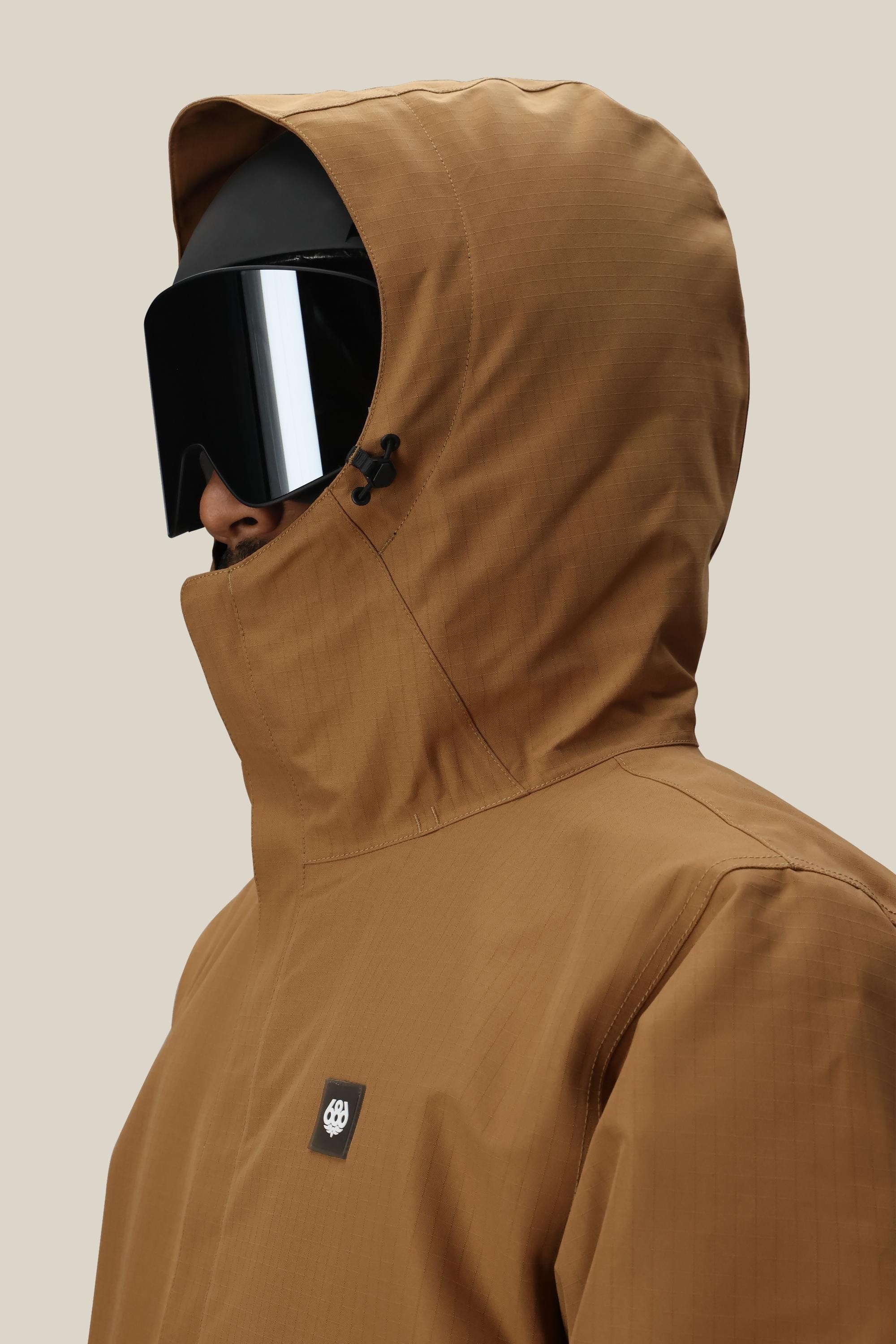 A person in a 686 Mens Foundation Insulated Jacket, paired with large black ski goggles and a black helmet, is photographed from the side against a plain beige background.