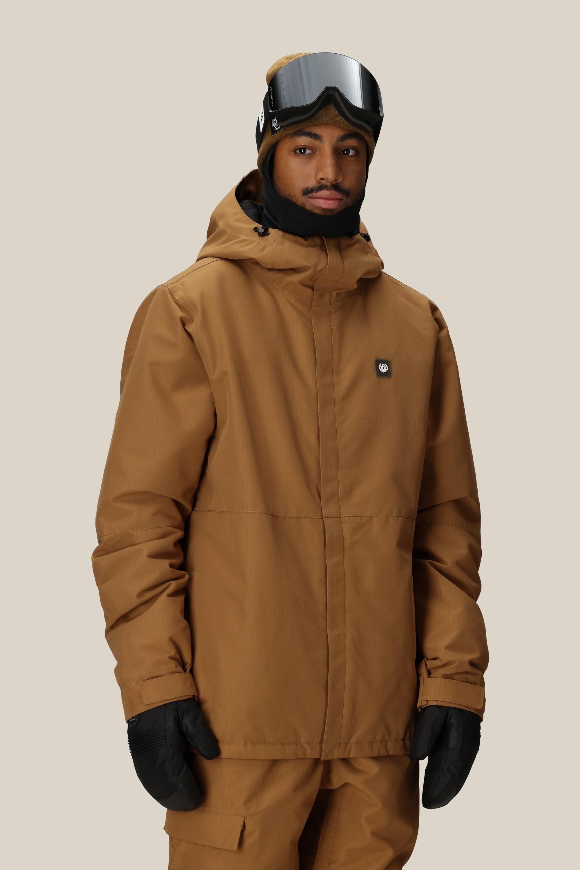 A person wearing the 686 Mens Foundation Insulated Jacket, featuring a brown hood, along with matching pants, black gloves, and ski goggles stands facing the camera against a light beige background.