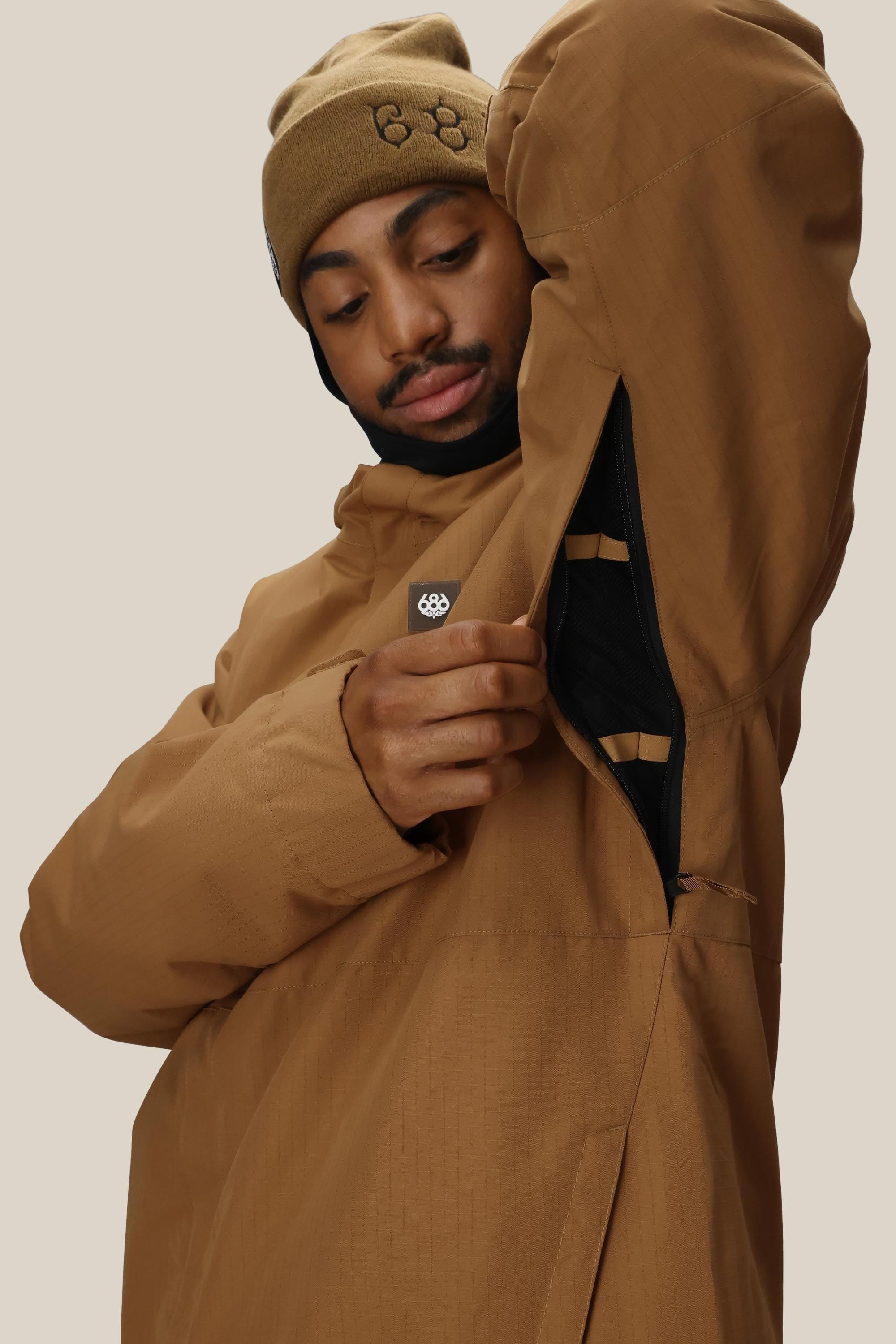 A man in a 686 Mens Foundation Insulated Jacket and beanie opens an underarm zip vent, revealing black lining. The jacket is brown, waterproof, and the background is plain and light-colored.