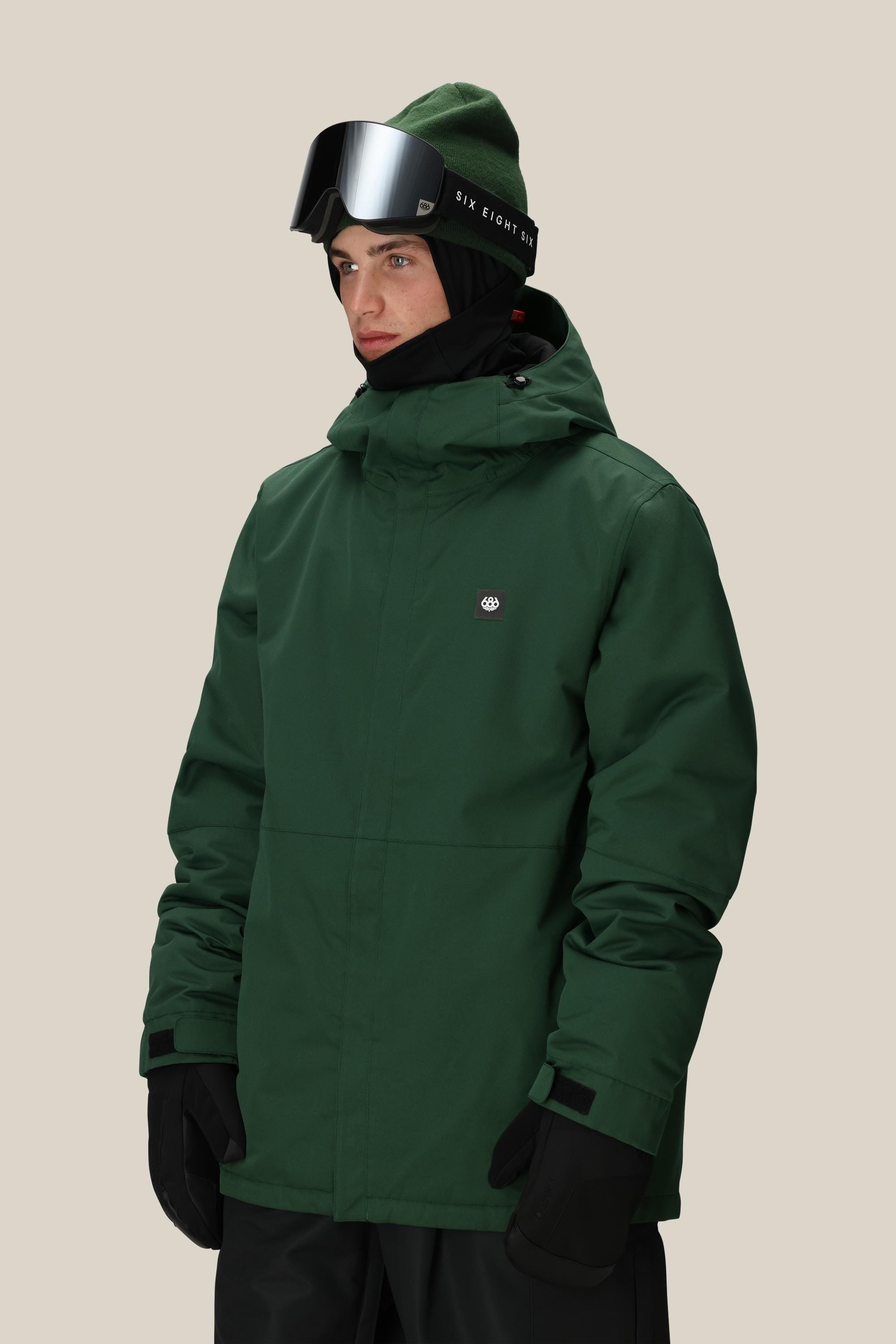 A person wearing the 686 Mens Foundation Insulated Jacket, black gloves, a green beanie, black ski goggles, and a black neck gaiter stands against a plain beige background.