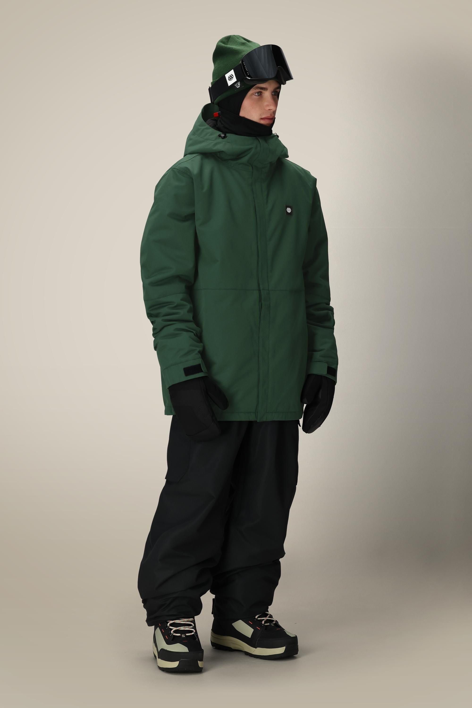 A person wearing a 686 Mens Foundation Insulated Jacket in green, black snow pants, black gloves, black boots, a black beanie, and ski goggles stands against a neutral background.