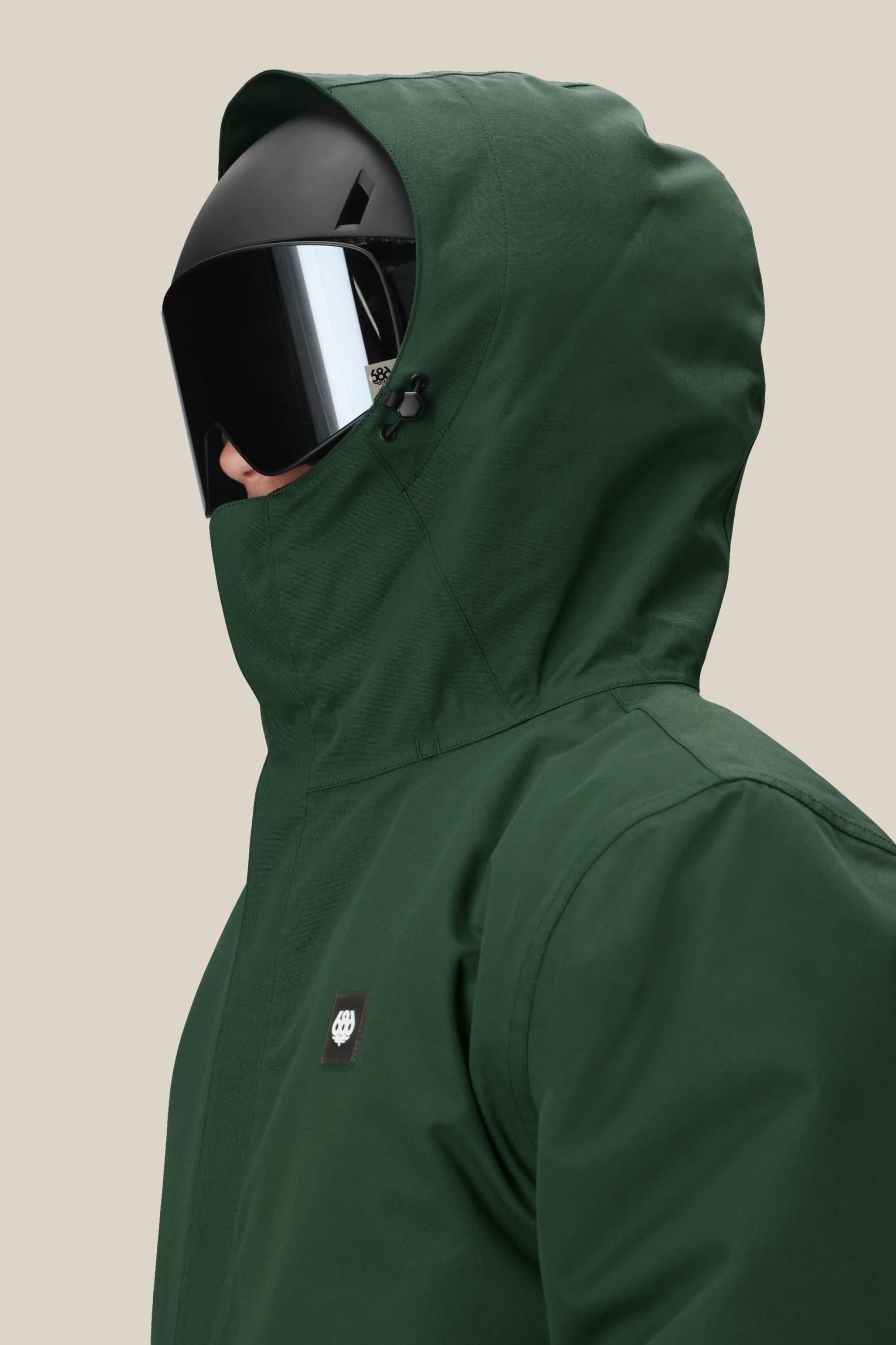 A person in a 686 Mens Foundation Insulated Jacket in dark green and a large black visor helmet, facing left against a plain beige background.