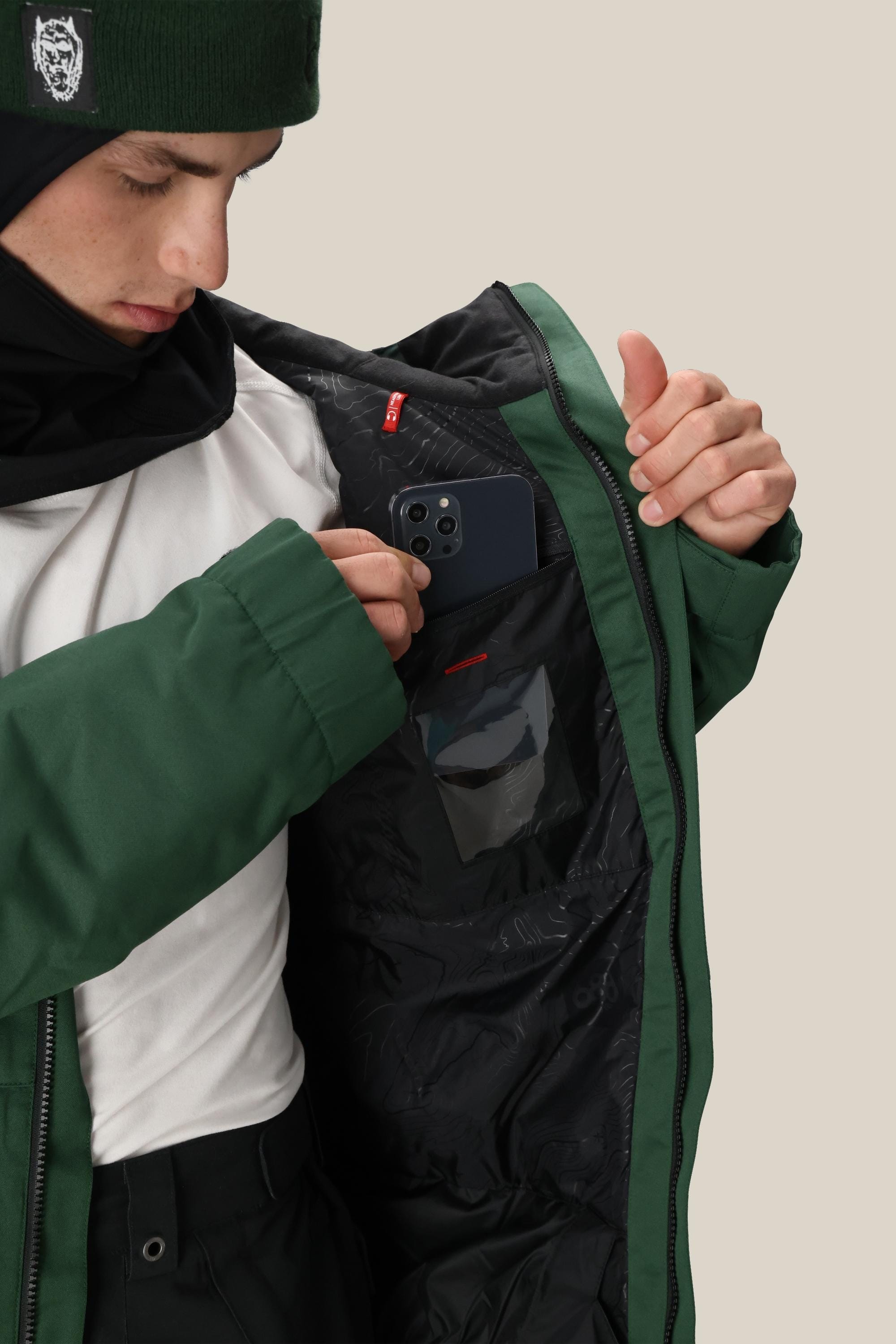 A man in a green 686 Mens Foundation Insulated Jacket and beanie places a smartphone into the jacket’s inner pocket, showcasing its black lining and pocket details against a plain beige background.