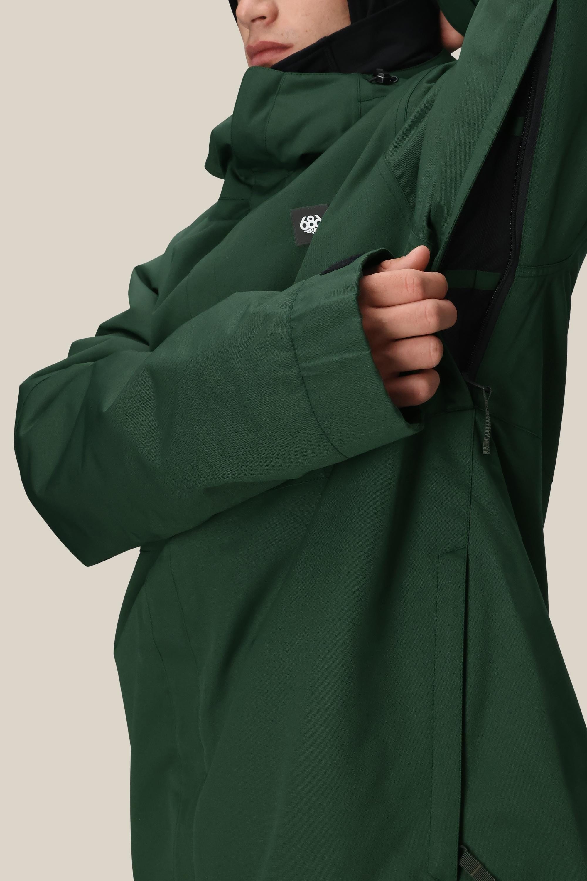 A person unzips a side vent under the arm of a green 686 Mens Foundation Insulated Jacket. The jacket features a high collar, small white geometric logo on the chest, and is set against a plain beige background.