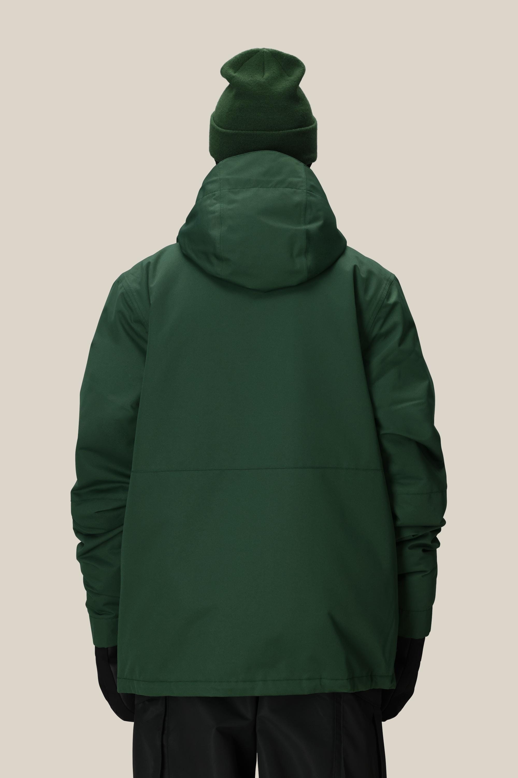 A person faces away from the camera, wearing a 686 Mens Foundation Insulated Jacket in dark green with a hood, paired with a green beanie, black gloves, and black pants, set against a plain light beige background.