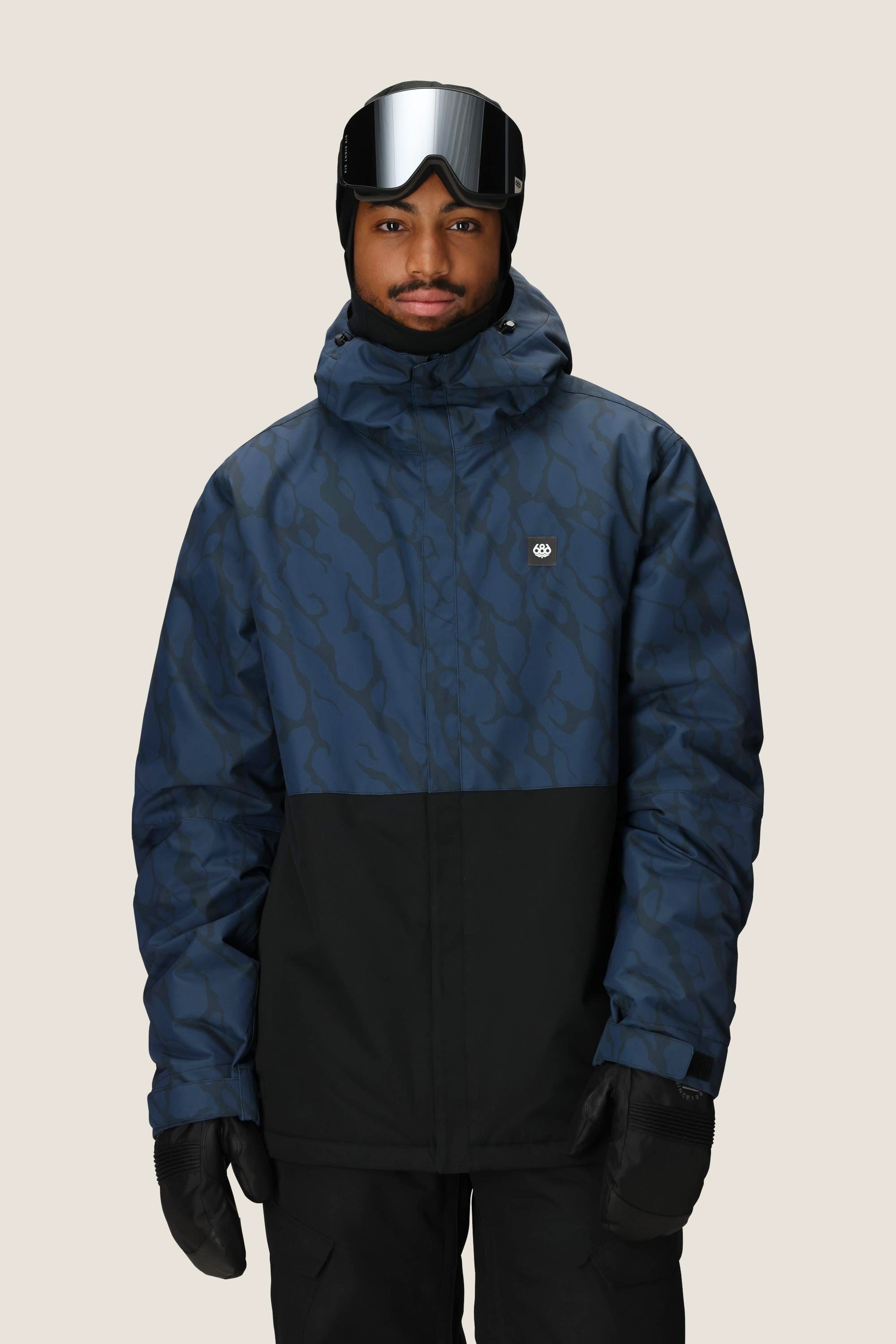 A person in a blue and black 686 Mens Foundation Insulated Jacket, black gloves, black beanie, and ski goggles stands facing forward against a plain light background.