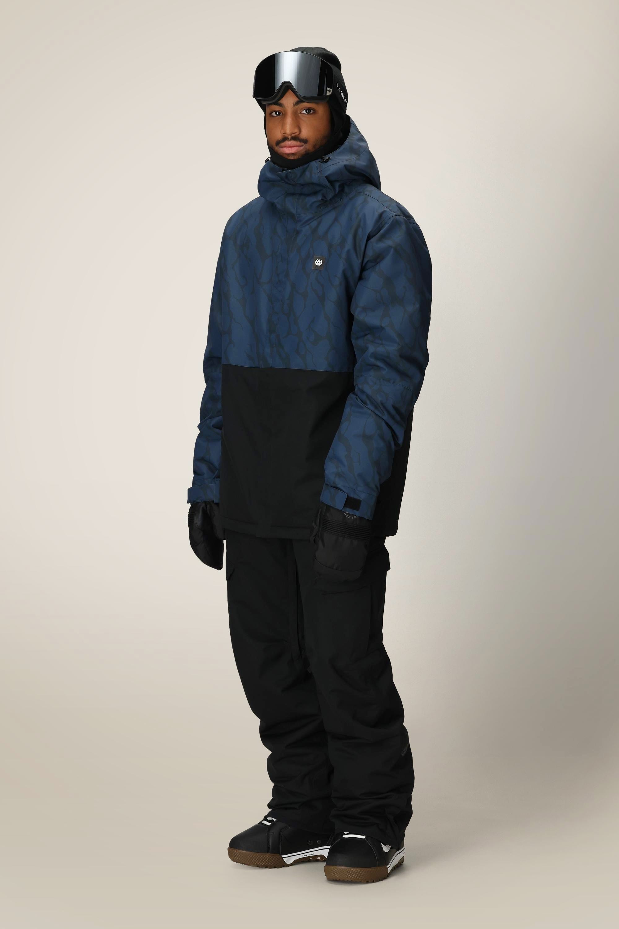 A man wearing the 686 Mens Foundation Insulated Jacket, black snow pants, gloves, dark snow boots, and ski goggles stands against a plain light background.