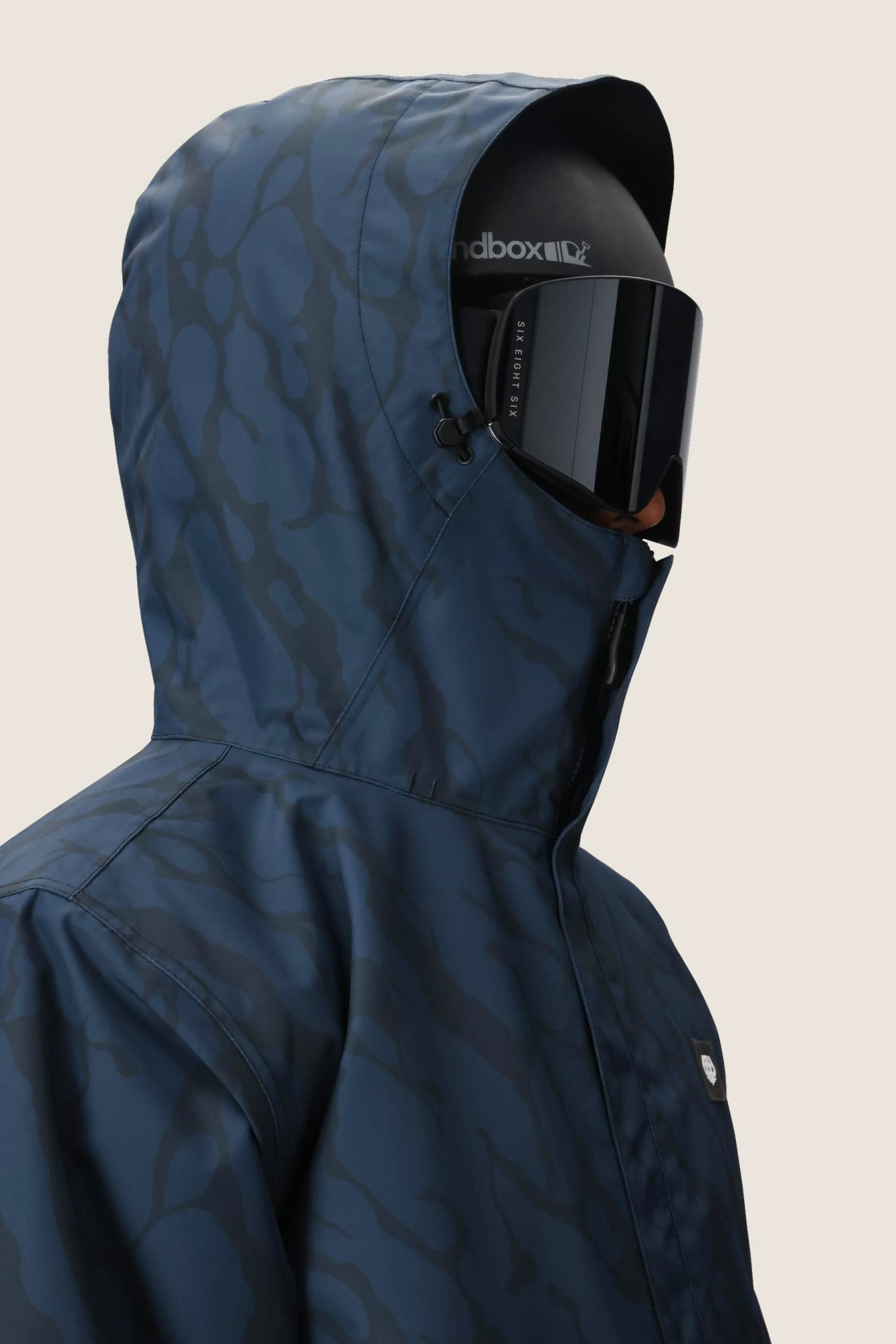 A person wearing the 686 Mens Foundation Insulated Jacket in blue camo, paired with black ski goggles and a helmet, is seen from the side against a plain background.