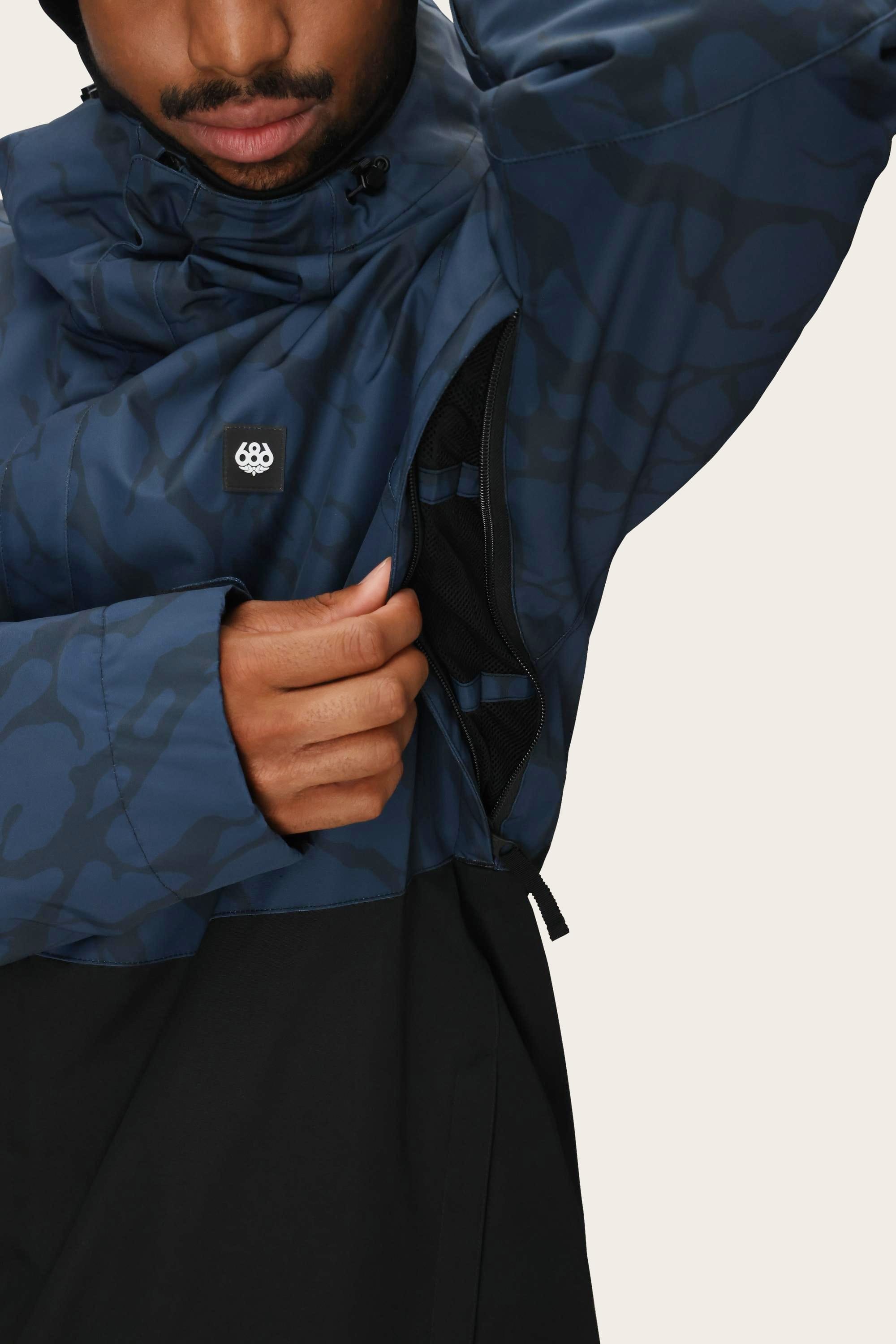 A person wearing the 686 Mens Foundation Insulated Jacket, a blue and black polyester waterproof jacket by 686, partially unzips a side vent to reveal its mesh lining. A small white logo appears on the chest.
