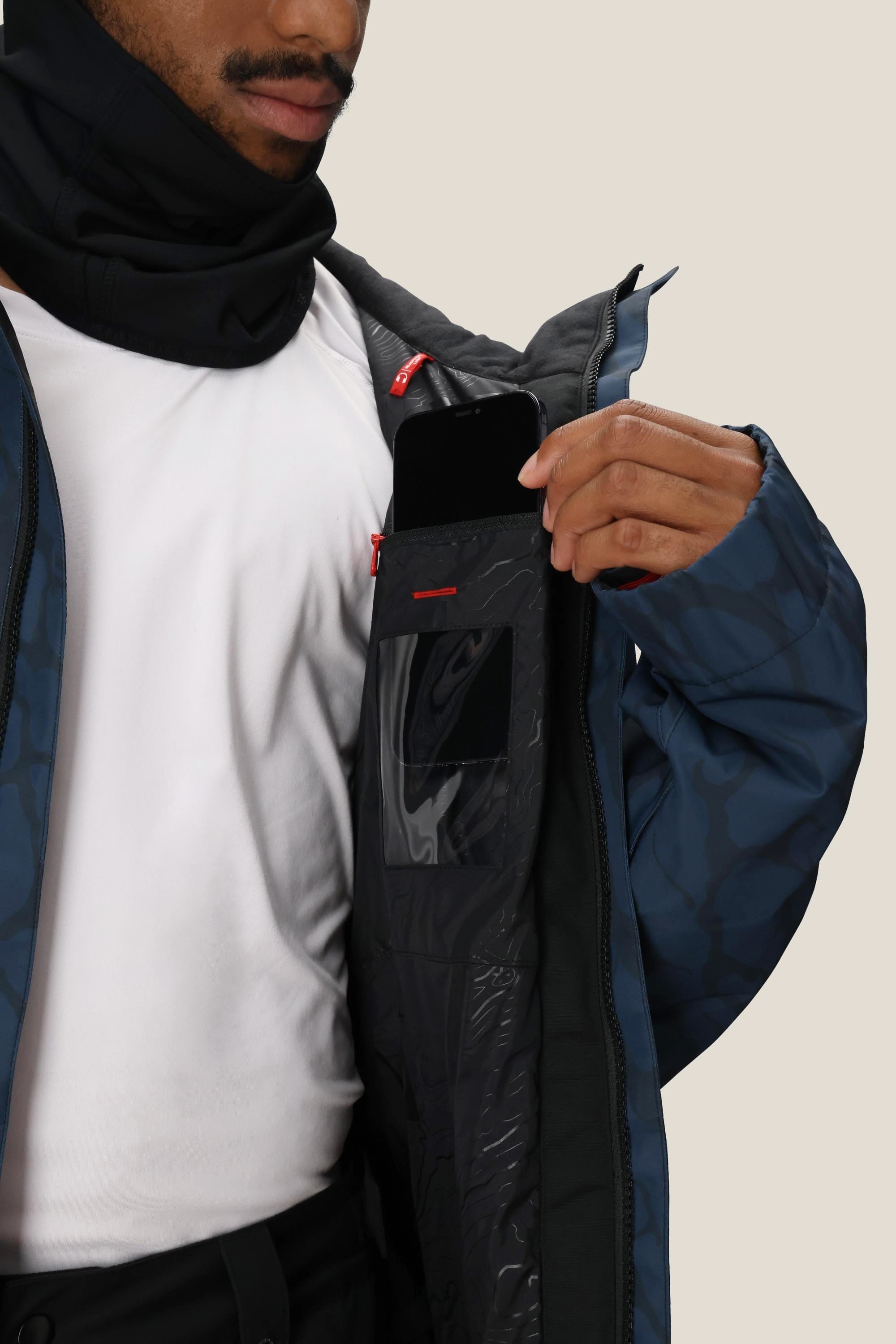 A man in a blue patterned 686 Mens Foundation Insulated Jacket and white shirt displays the jackets inside pocket, holding a black smartphone halfway into it.