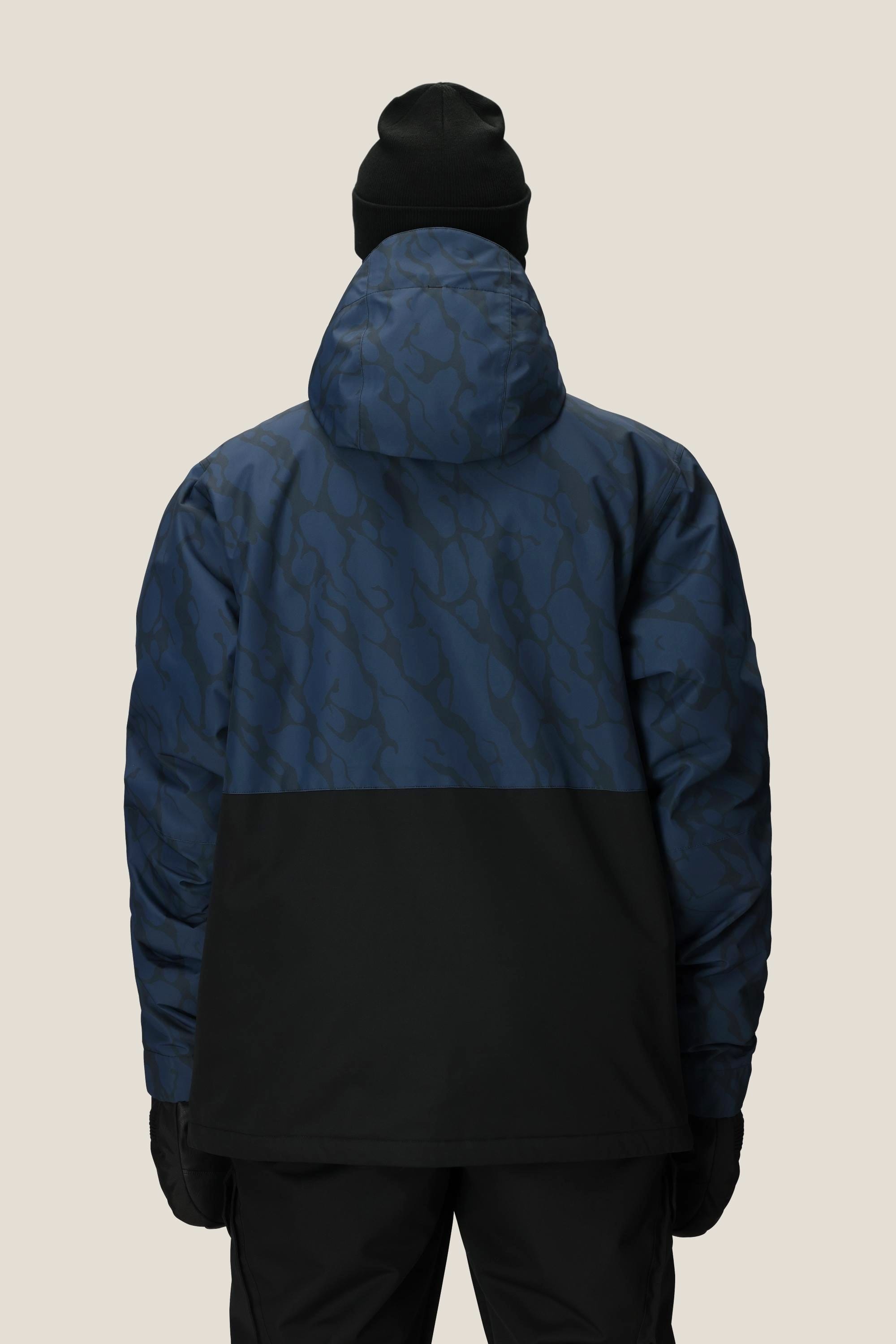 A person wearing a black beanie, gloves, and pants is shown from the back in the 686 Mens Foundation Insulated Jacket by 686, featuring a blue and black pattern, against a plain beige background.