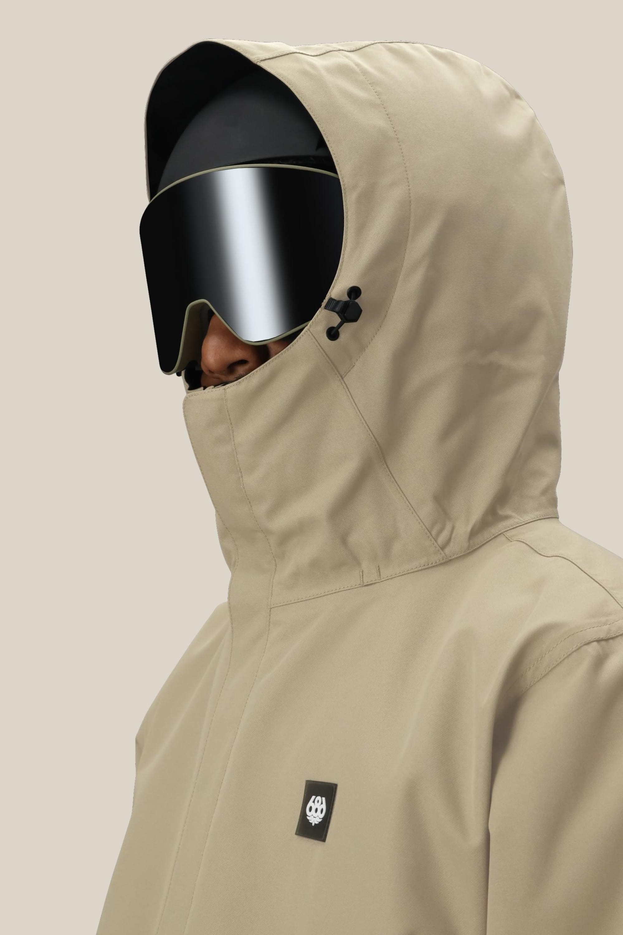 A person wears the 686 Mens Foundation Insulated Jacket in beige with a hood, black ski goggles, and a black helmet, their face mostly hidden. The plain light beige background highlights the jacket’s design by 686.