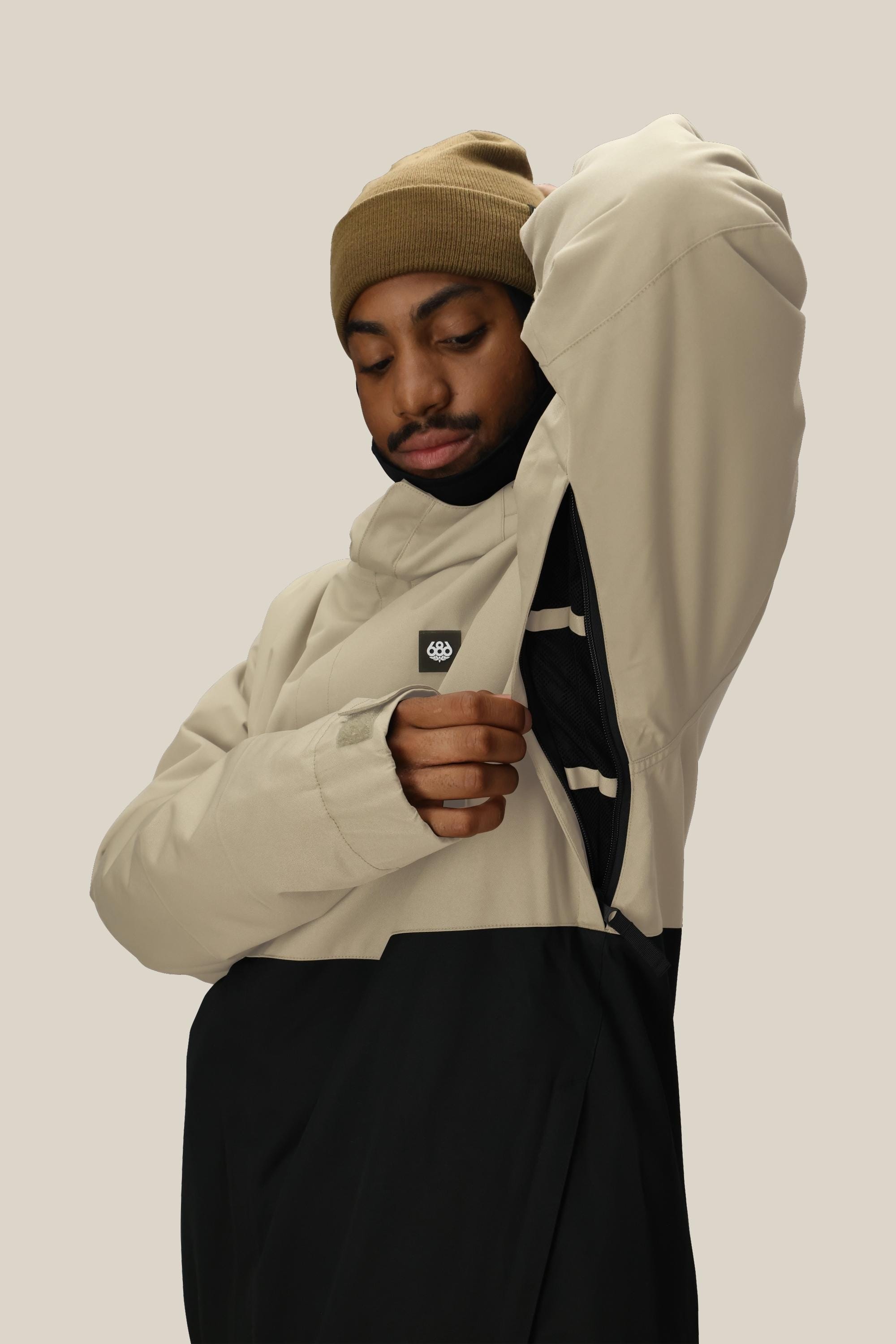 A person wearing a tan beanie and the 686 Mens Foundation Insulated Jacket in beige and black lifts their arm to show the side zipper. The background is plain and light-colored.