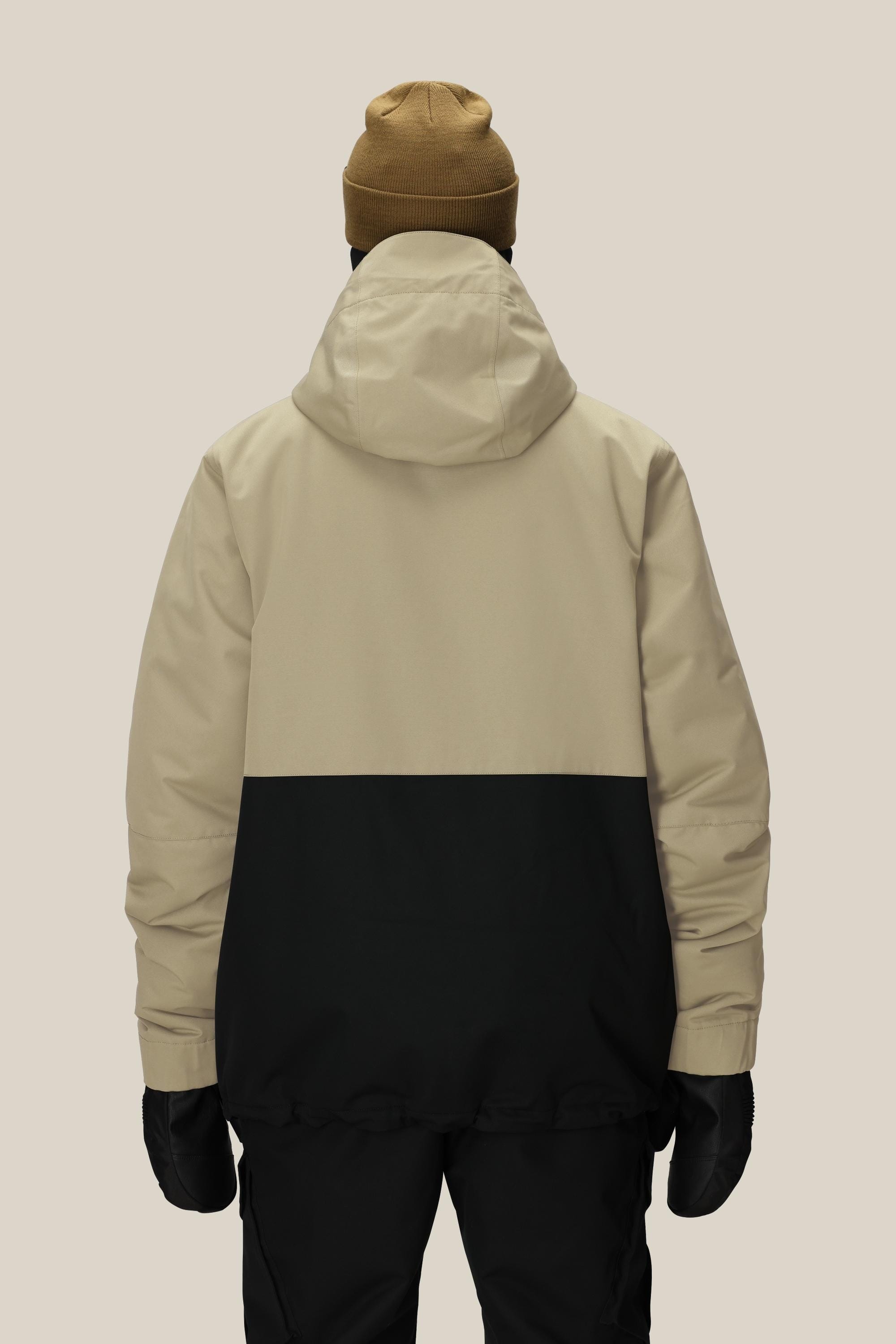 A person wearing the 686 Mens Foundation Insulated Jacket in tan and black with a hood, paired with black pants, black gloves, and a brown beanie, is photographed from behind against a plain beige background.