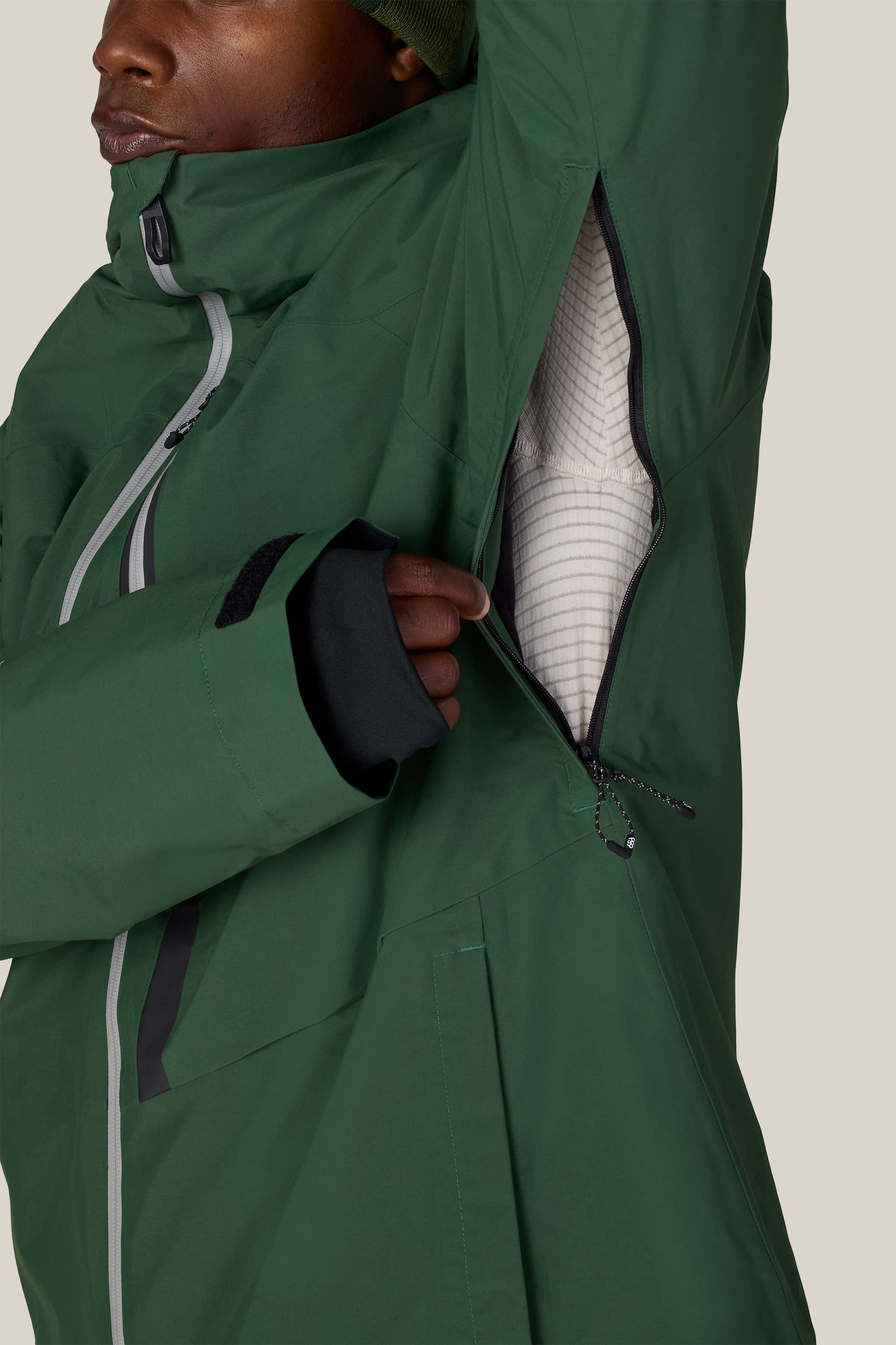 A person wearing the 686 Men’s GORE-TEX® GT Thermagraph® Jacket in green unzips an underarm vent, revealing a white inner lining. The jacket by 686 features black details and Velcro straps on the sleeves.