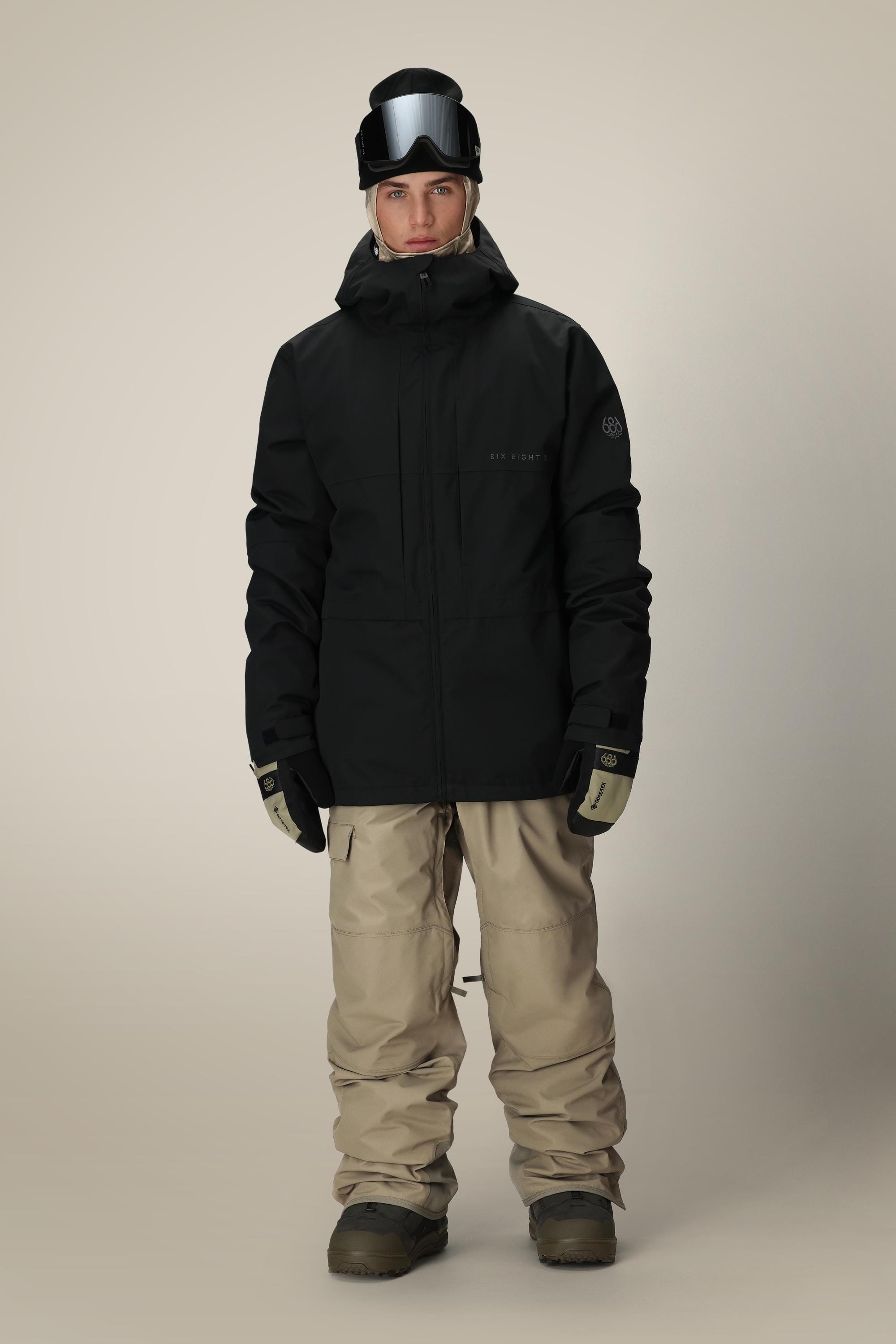 A person faces forward, wearing the 686 Mens Smarty 3-In-1 Form Jacket in black with a hood, plus beige snow pants, black gloves, brown snow boots, and a black helmet with ski goggles lifted above their eyes. The background is plain and light.