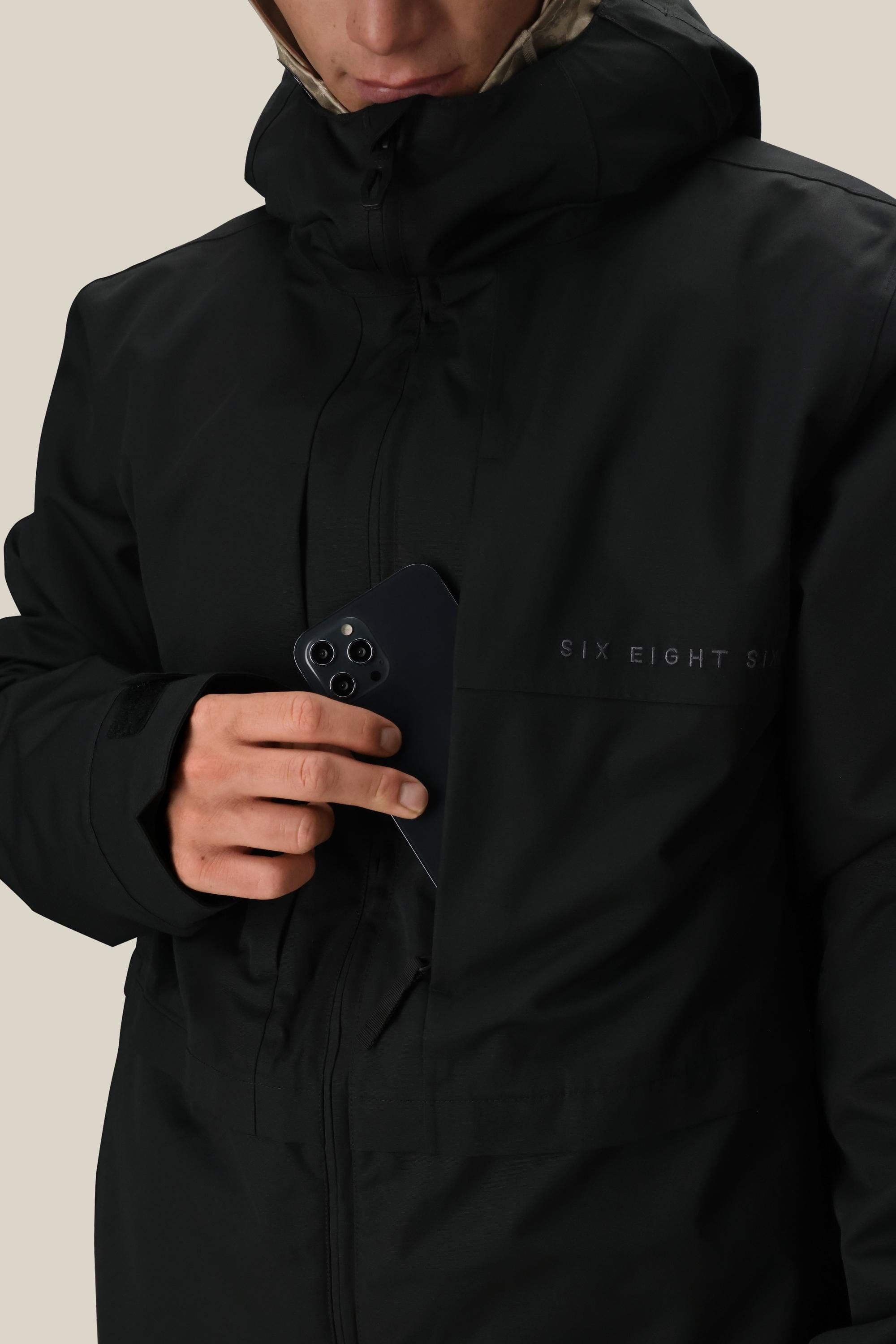 A person in a 686 Mens Smarty 3-In-1 Form Jacket places a black smartphone into the chest pocket while facing slightly downward, displaying the 686 logo.
