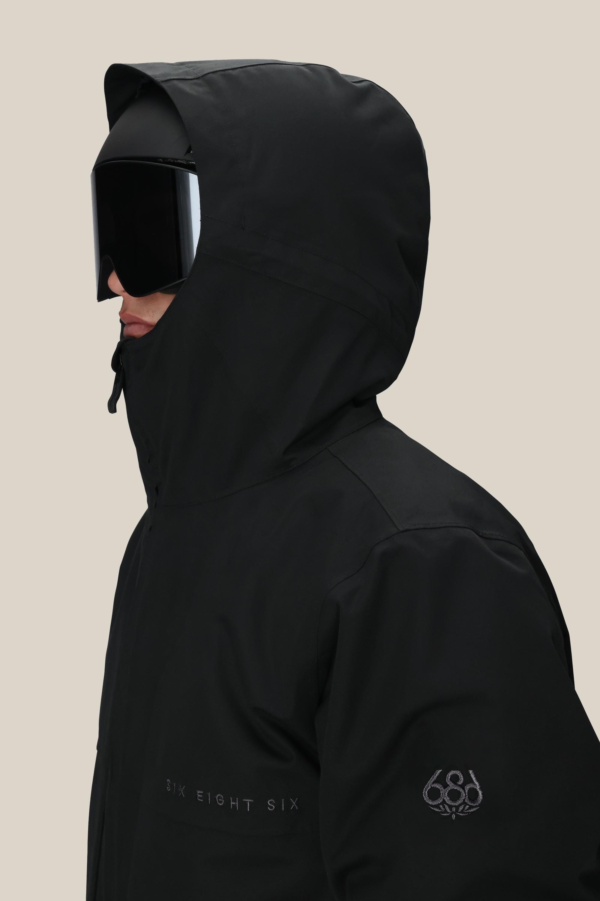 A person in a black 686 Mens Smarty 3-In-1 Form Jacket with 686 on the sleeve and chest, wearing large black ski goggles, shown from the side against a beige background.