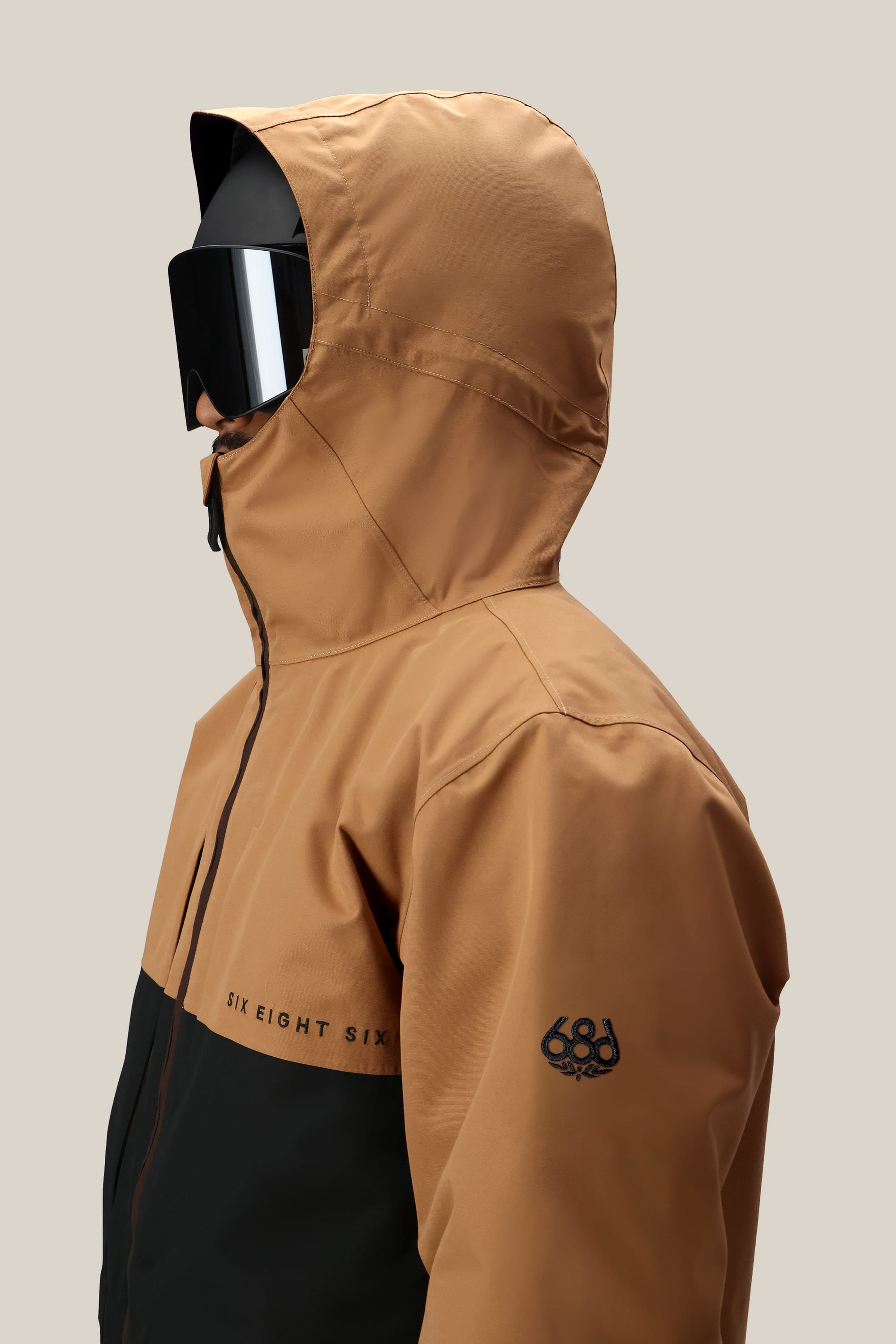 A person wearing the 686 Mens Smarty 3-In-1 Form Jacket in brown and black, featuring the 686 logo on the sleeve, along with black ski goggles. The background is plain and light-colored.