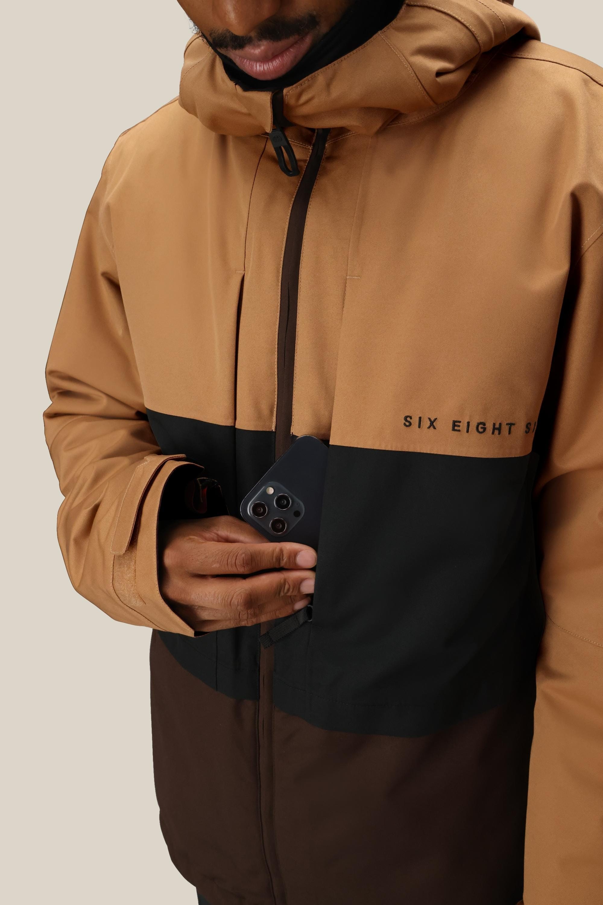 A person in a brown and black 686 Mens Smarty 3-In-1 Form Jacket is putting a black smartphone into the inner chest pocket. The label 686 is visible, their face is partially shown, and the background is plain beige.