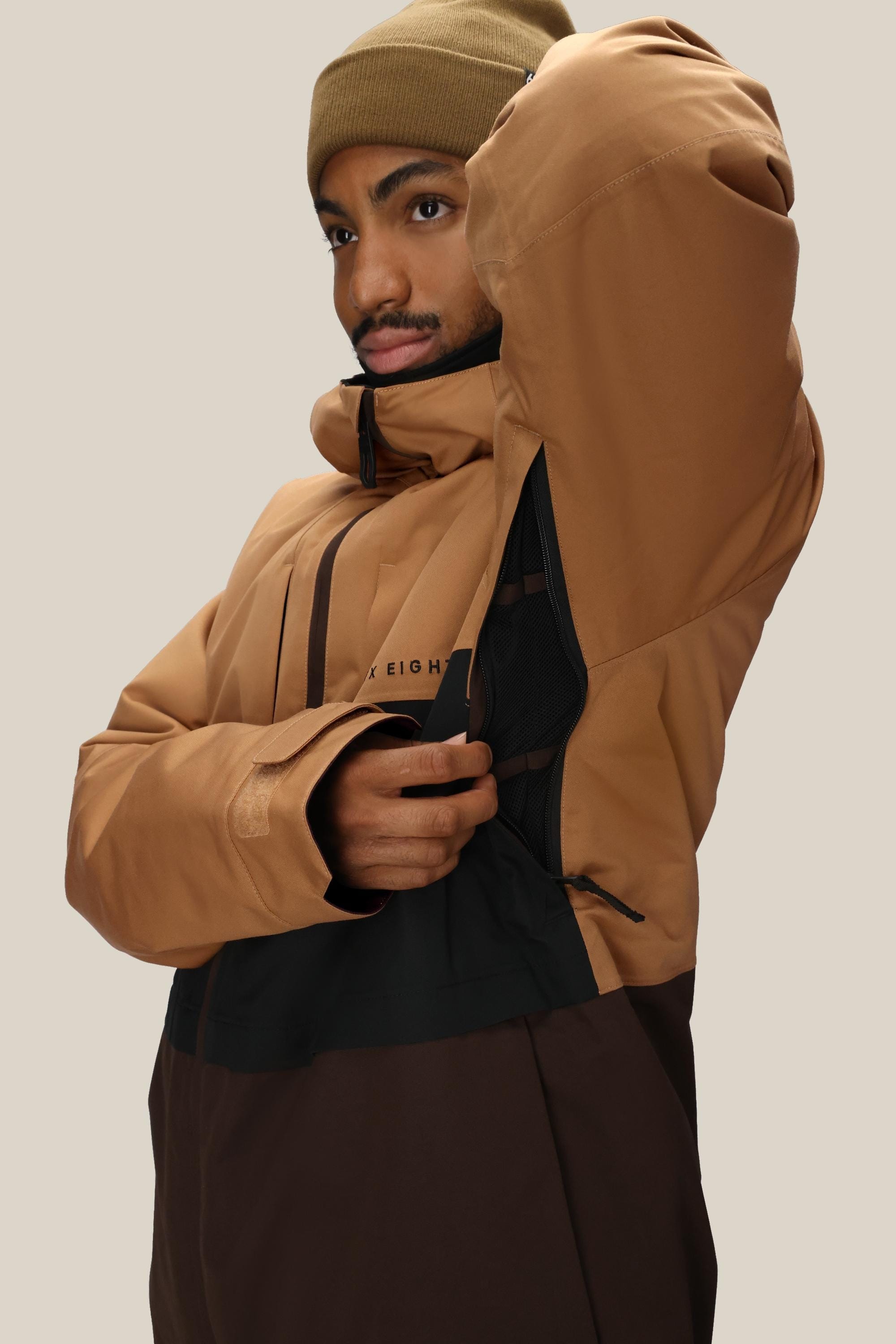 A person wearing a 686 Mens Smarty 3-In-1 Form Jacket in brown and tan with a tan beanie unzips the side vent, showing the black lining. The background is plain and light-colored.